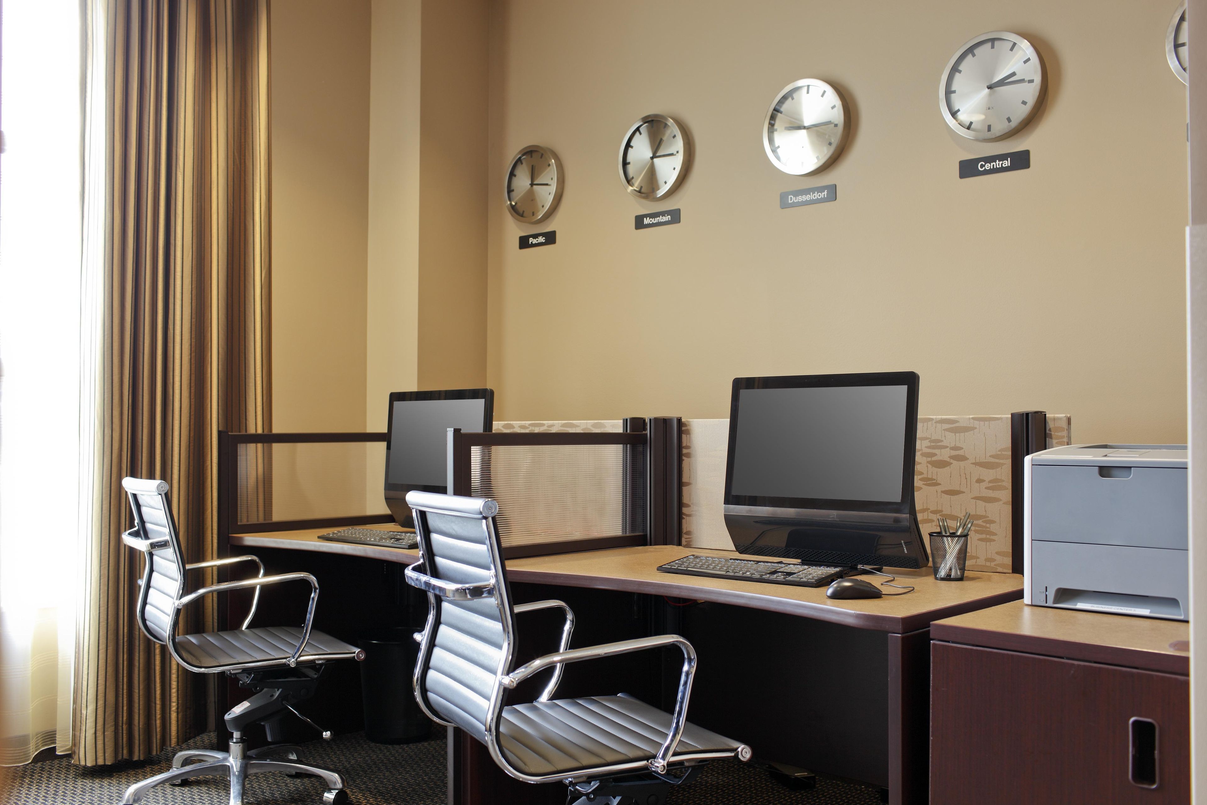 Hotelkamer met een bureau, twee stoelen, een computer, een printer en vier klokken met verschillende locaties.