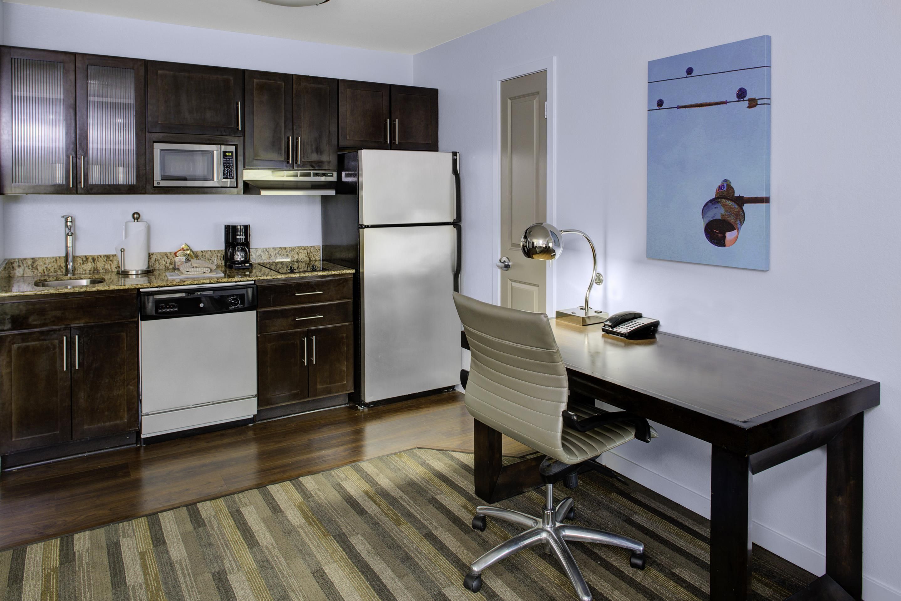 A compact kitchen with dark cabinetry, a microwave, a refrigerator, a coffee maker, and a dining area with a desk and chair.