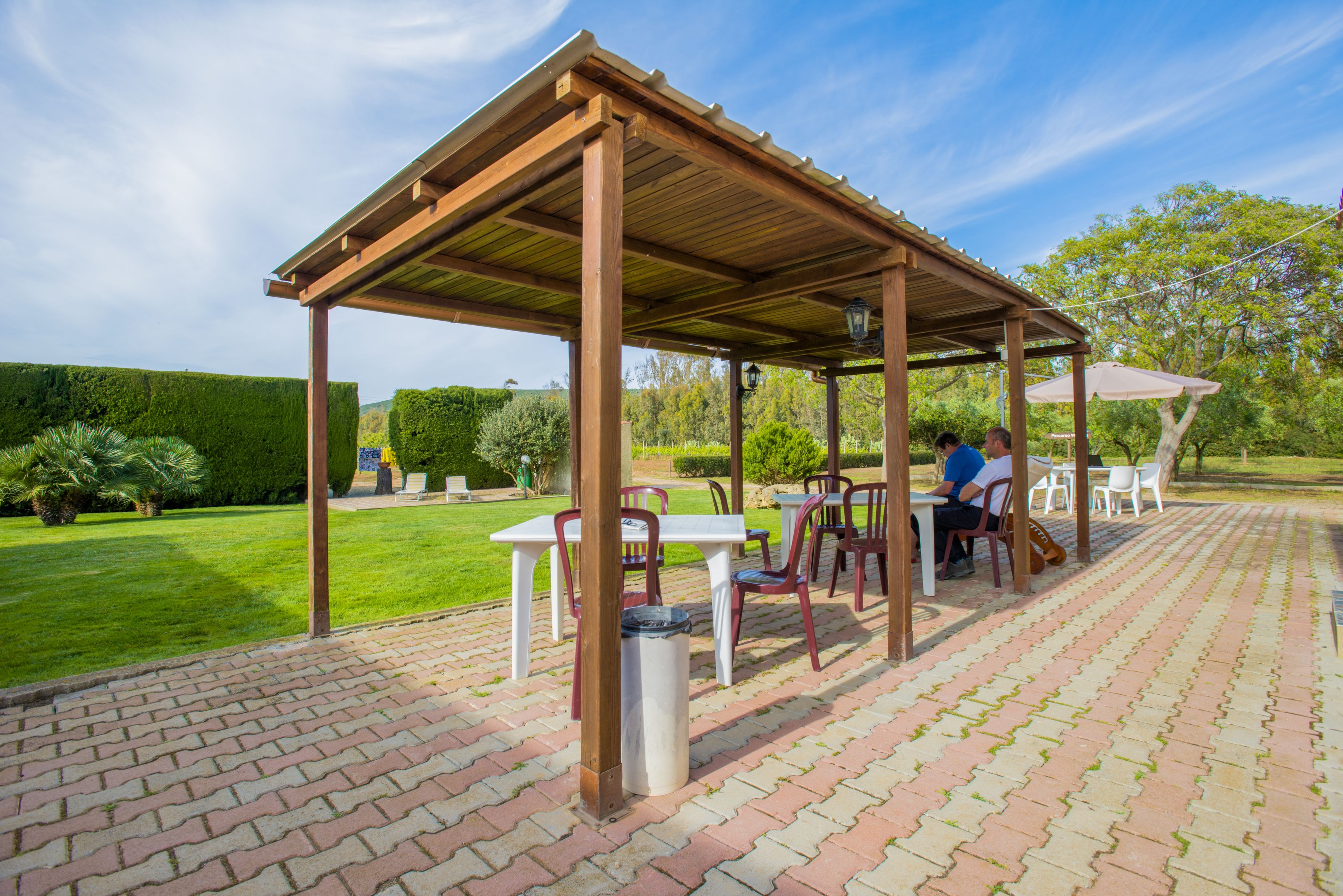 A covered outdoor seating area with tables and chairs, surrounded by a brick patio and greenery.