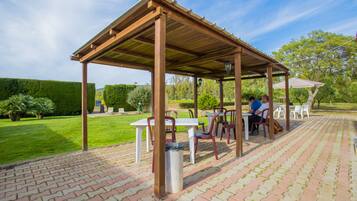 A covered outdoor seating area with tables and chairs, surrounded by a brick patio and greenery.