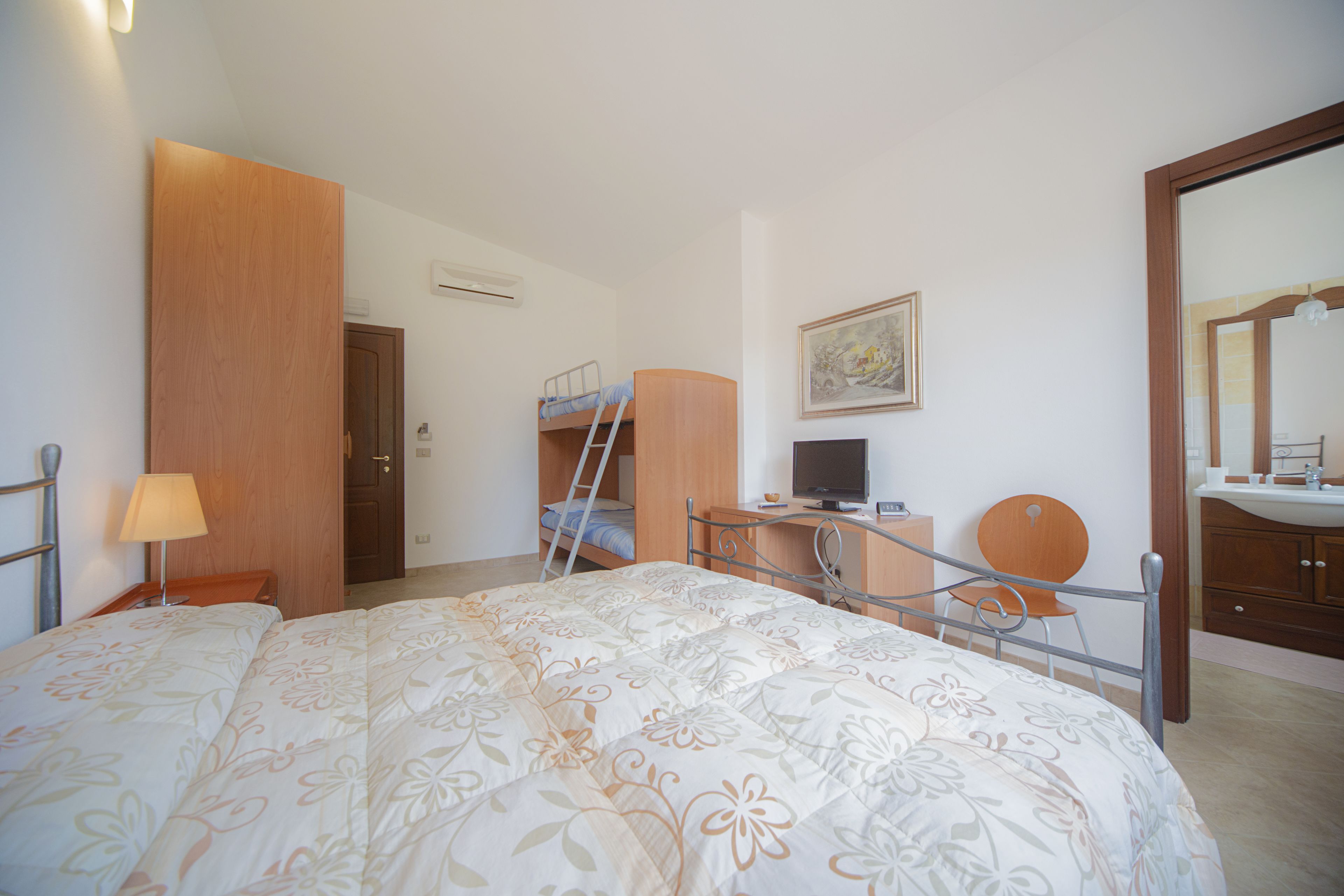 Una camera da letto con un letto a castello, una scrivania con sedia, una televisione e un bagno con lavandino e specchio.