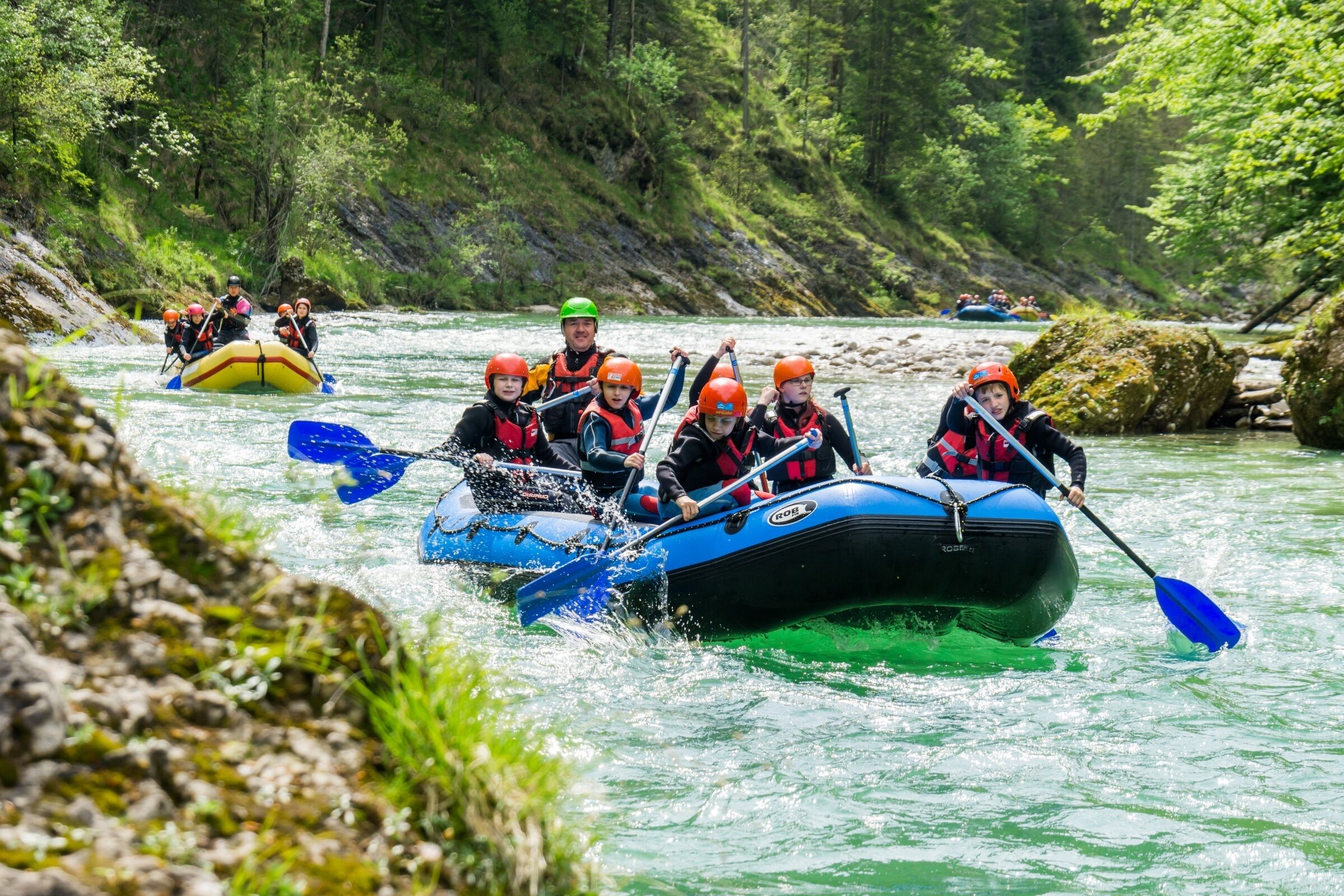 A group of people in a blue inflatable raft navigating a river surrounded by greenery.