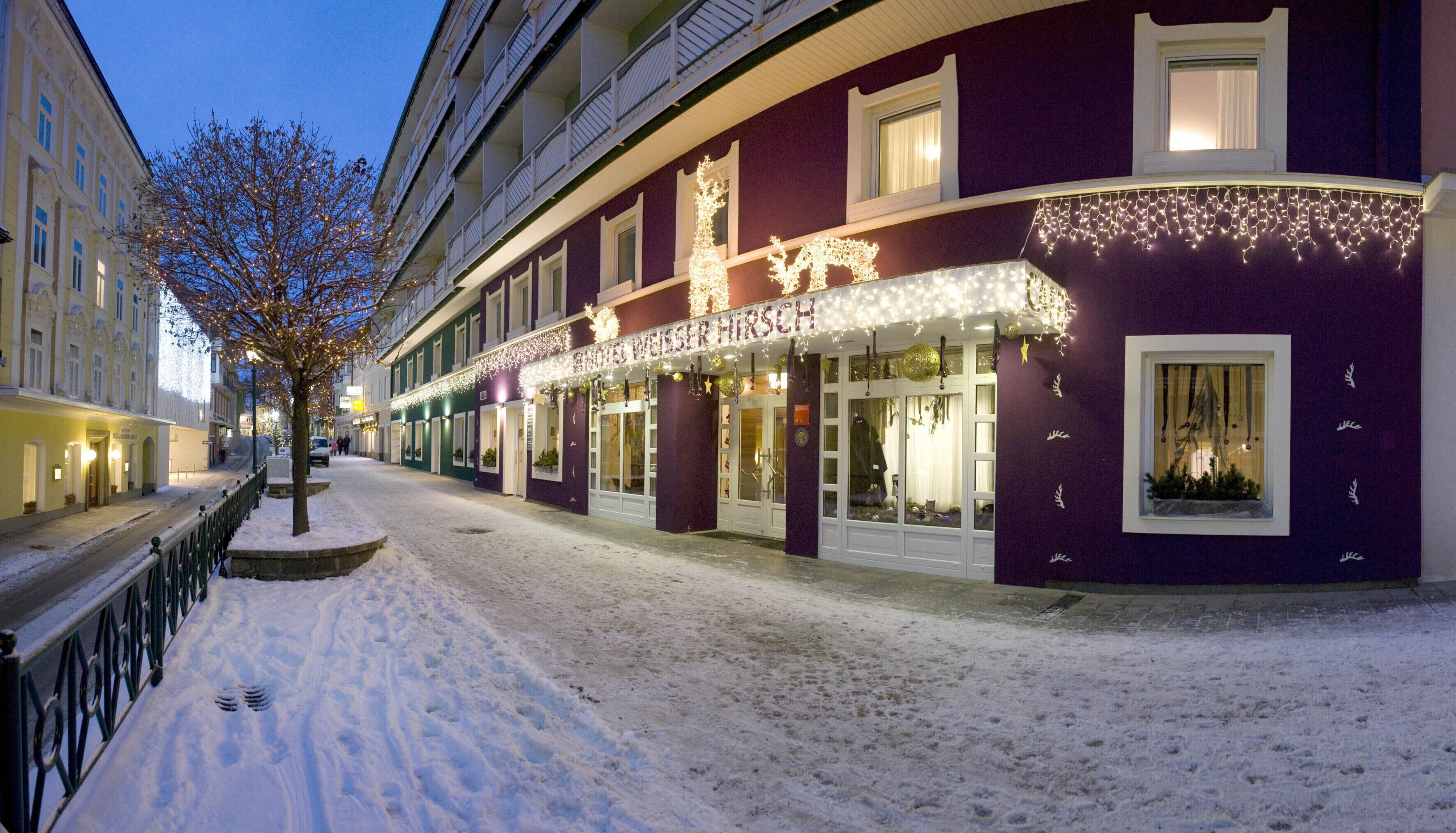 Um hotel com uma rua coberta de neve, uma árvore com luzes e fachada roxa.