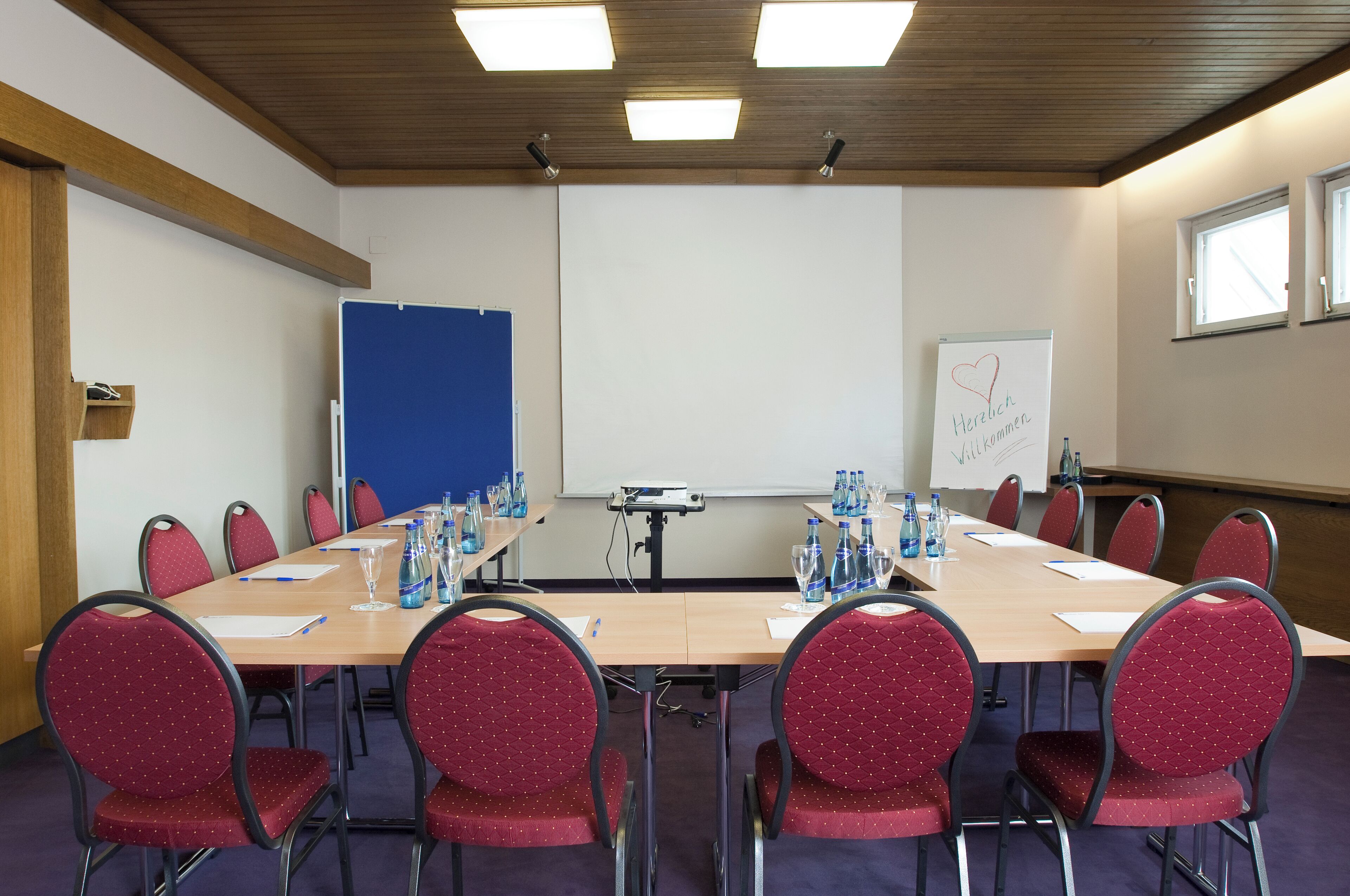 Une salle de conférence avec une longue table, des chaises rouges, un écran de projection et un tableau blanc.