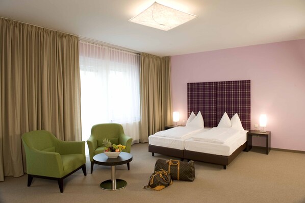 A hotel room with a bed, two armchairs, a small table with a fruit bowl, and a suitcase on the floor.