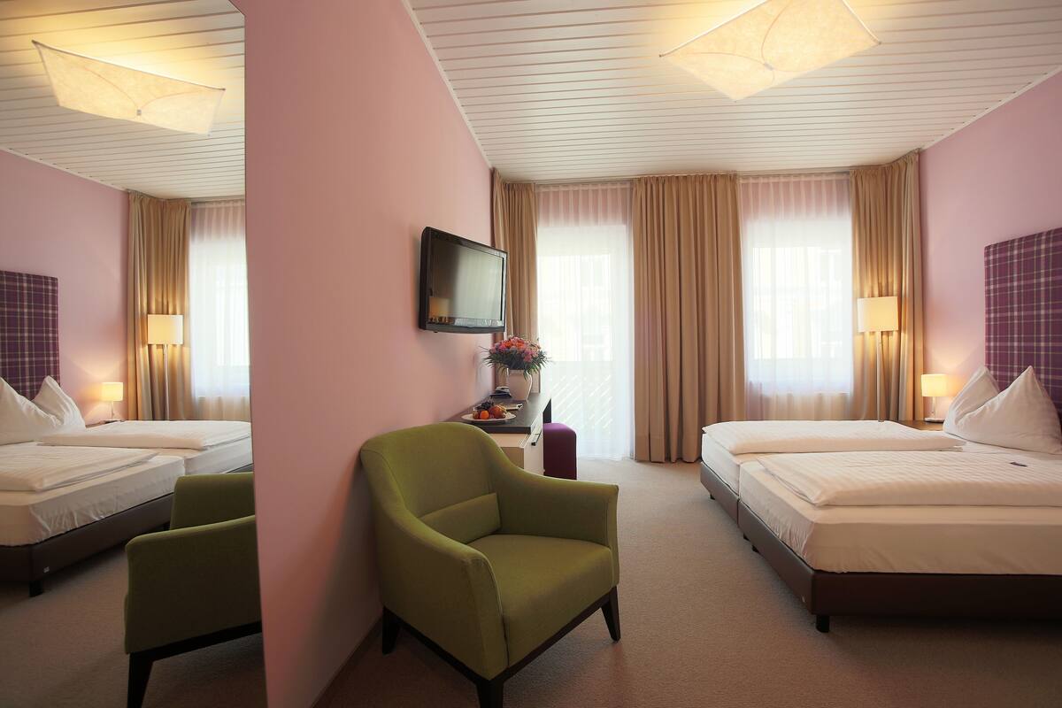 A hotel room with two beds, a green armchair, a flat-screen TV, and a desk with a lamp and a vase of flowers.