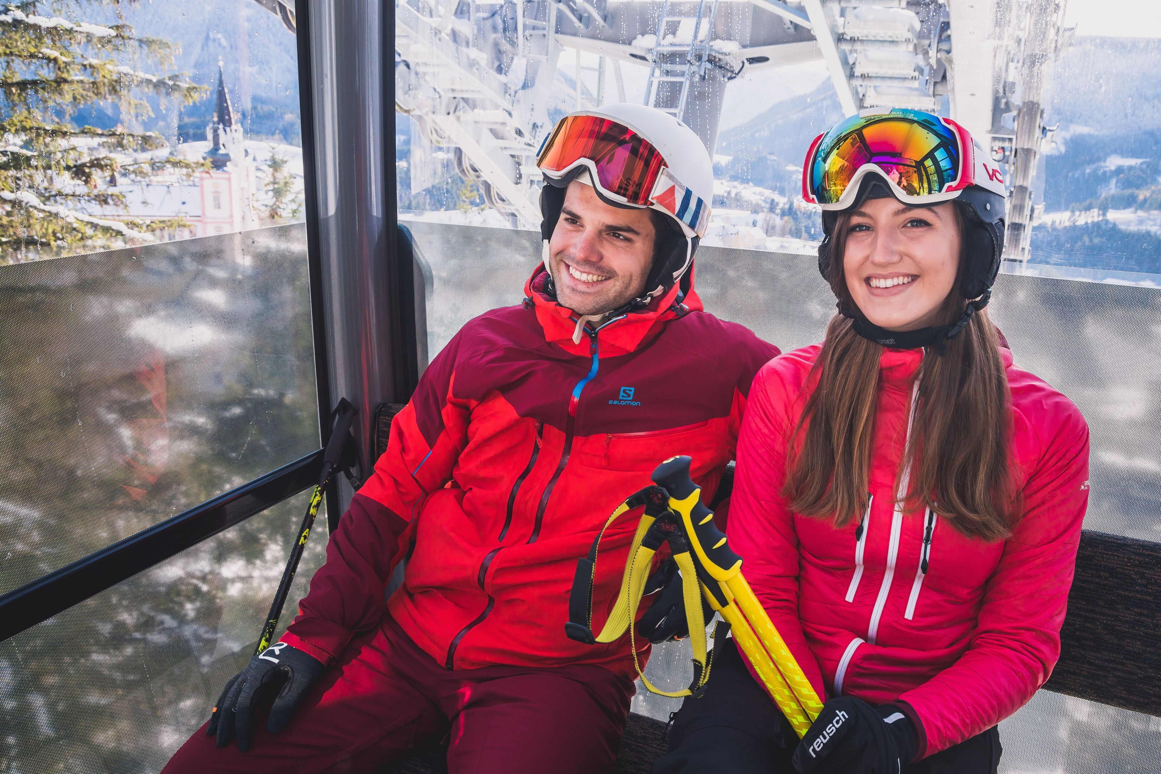 Two people in red ski gear sitting inside a vehicle with a view of a snowy mountain landscape.