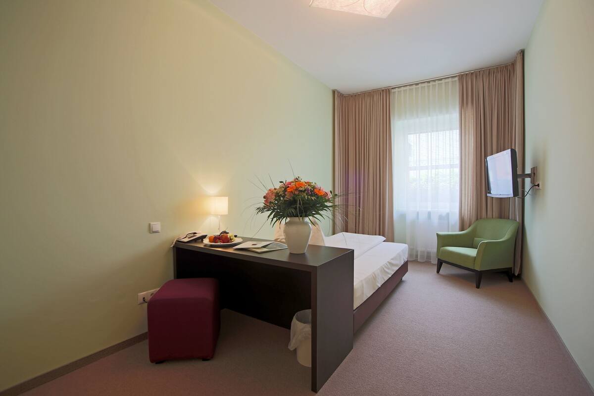 A hotel room with a bed, a desk with a lamp, a chair, and a TV mounted on the wall.