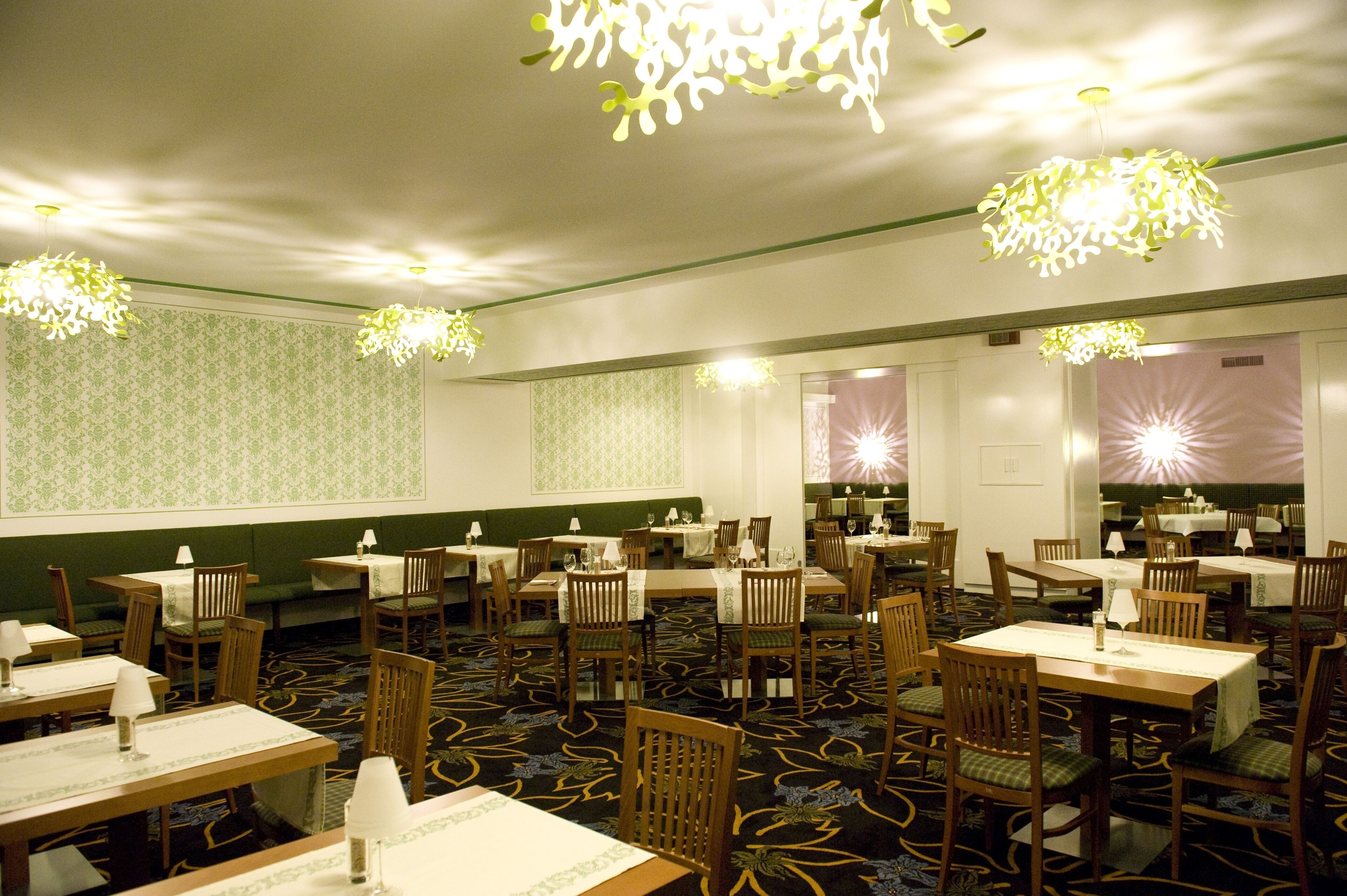 Un comedor con varias mesas y sillas, papel tapiz estampado en verde y candelabros florales colgantes.