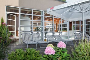 Un área de estar moderna al aire libre con sillones reclinables y una sombrilla, rodeada de follaje y plantas en macetas.