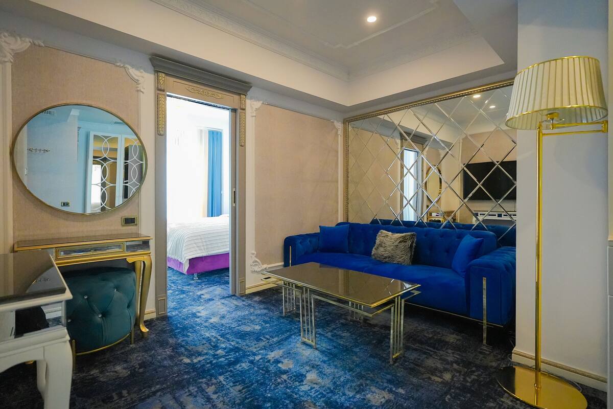 Una habitación de hotel con un sofá azul, un espejo redondo, una mesa de centro de vidrio y una lámpara de pie.