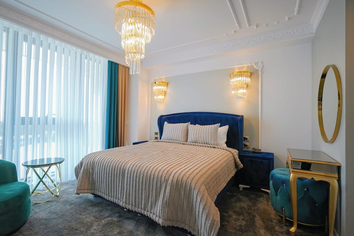 Habitación de hotel con una cama grande, una mesita de noche, una pequeña mesa redonda, un sillón verde y un reposapiés azul.