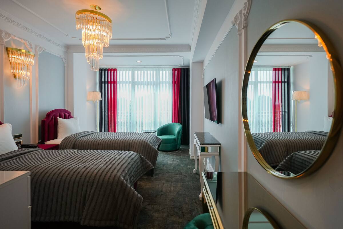Habitación de hotel con dos camas, un sillón verde, un televisor y un espejo grande.