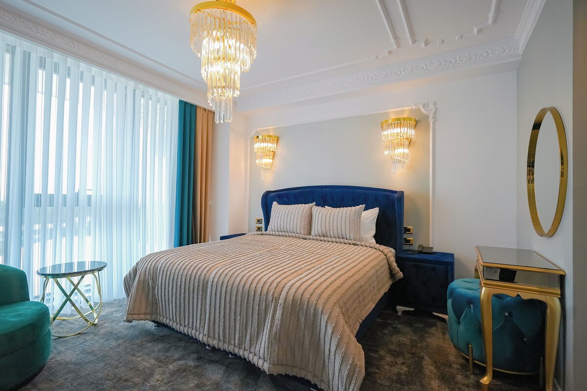 Una camera d'albergo con un letto grande, un comodino, un tavolino rotondo, una poltrona verde e un poggiapiedi blu.