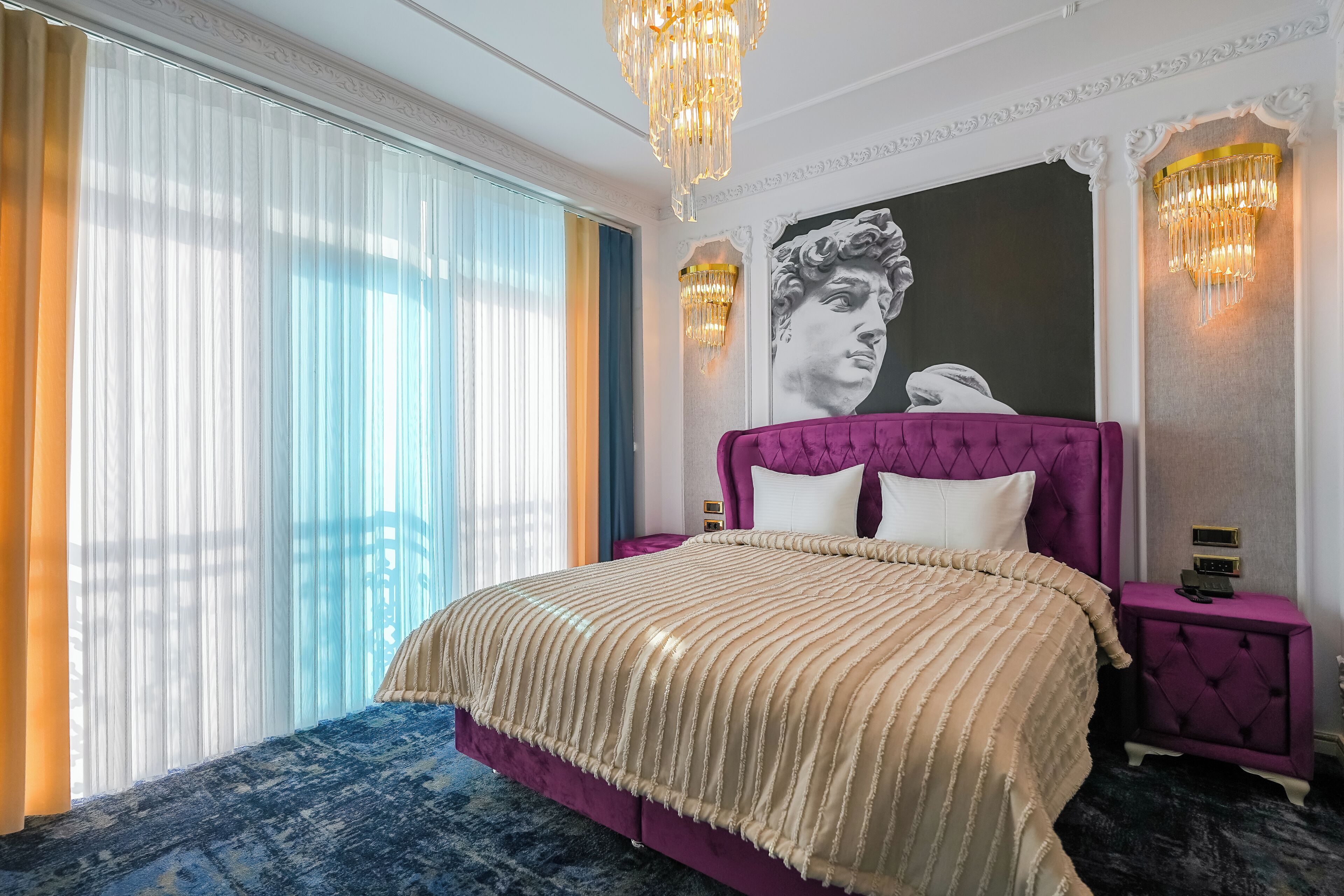 Une chambre à coucher avec un grand lit, une tête de lit violette et une grande fenêtre ornée de rideaux légers.