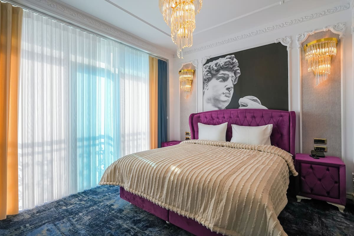Un dormitorio con una cama grande, un cabecero morado y un ventanal con cortinas translúcidas.