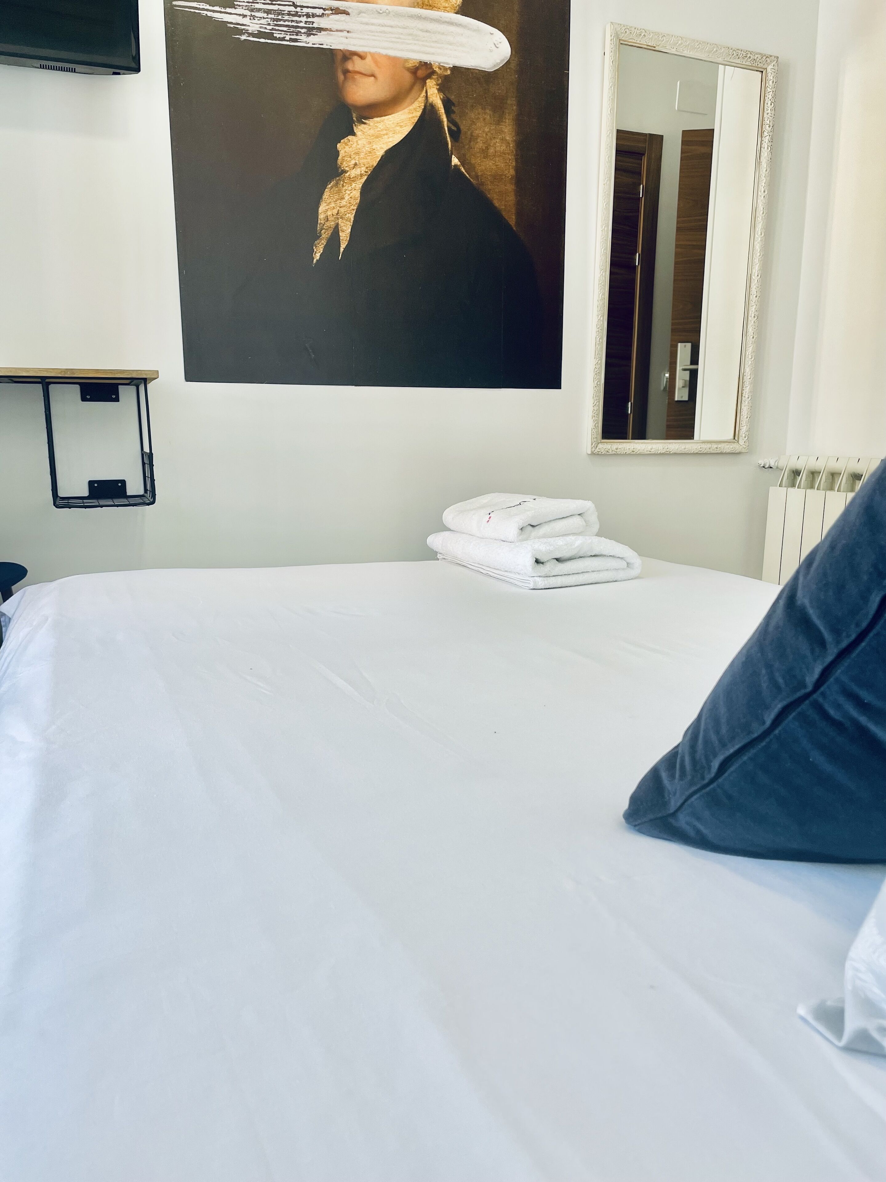 A hotel room with a neatly made bed, a stack of white towels, a large mirror, and a painting on the wall.