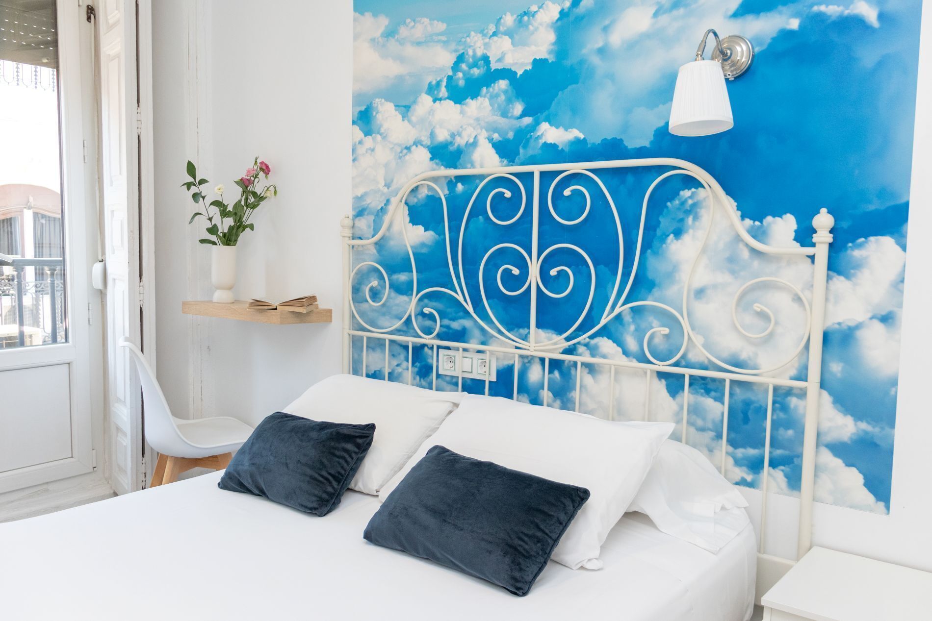 A bedroom with a bed, pillows, a nightstand, a wall-mounted lamp, and a decorative wall with a sky and clouds theme.
