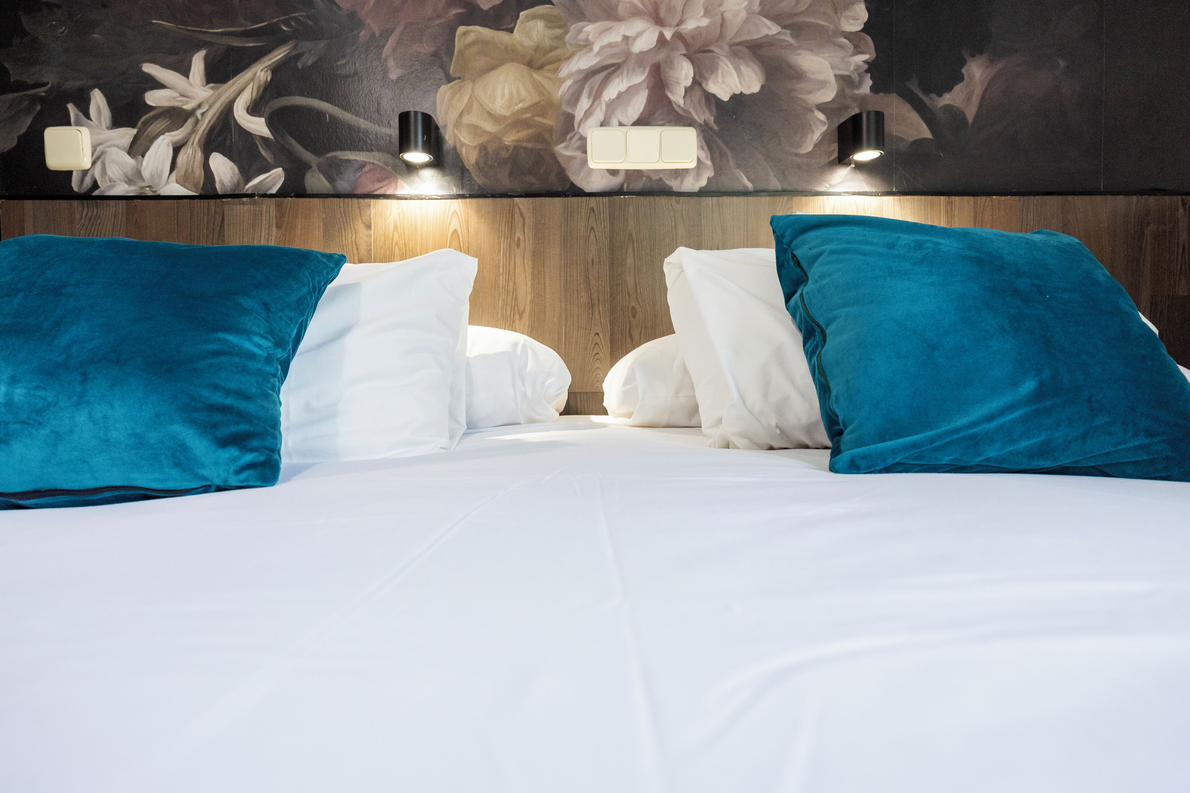 A bed with white linens and teal pillows, a wooden headboard, and a floral mural on the wall.