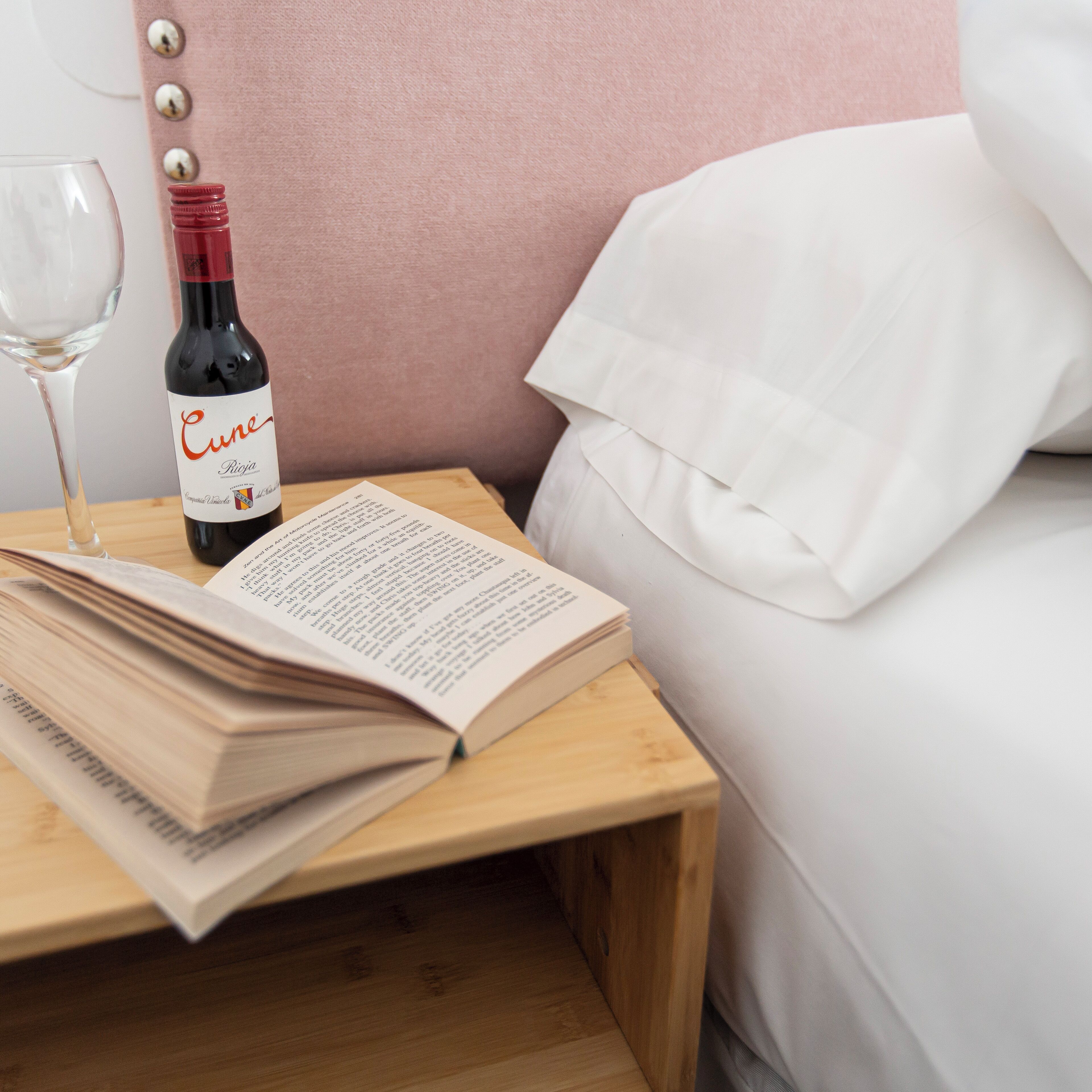 Una mesita de noche de madera con una botella de vino, una copa y un libro abierto.
