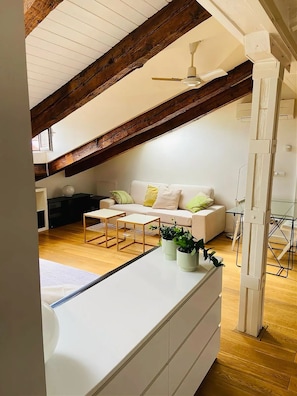 A loft-style room with a high ceiling, exposed wooden beams, a white sofa set, two small wooden tables, and a ceiling fan. - Hostal Veracruz (Madrid)