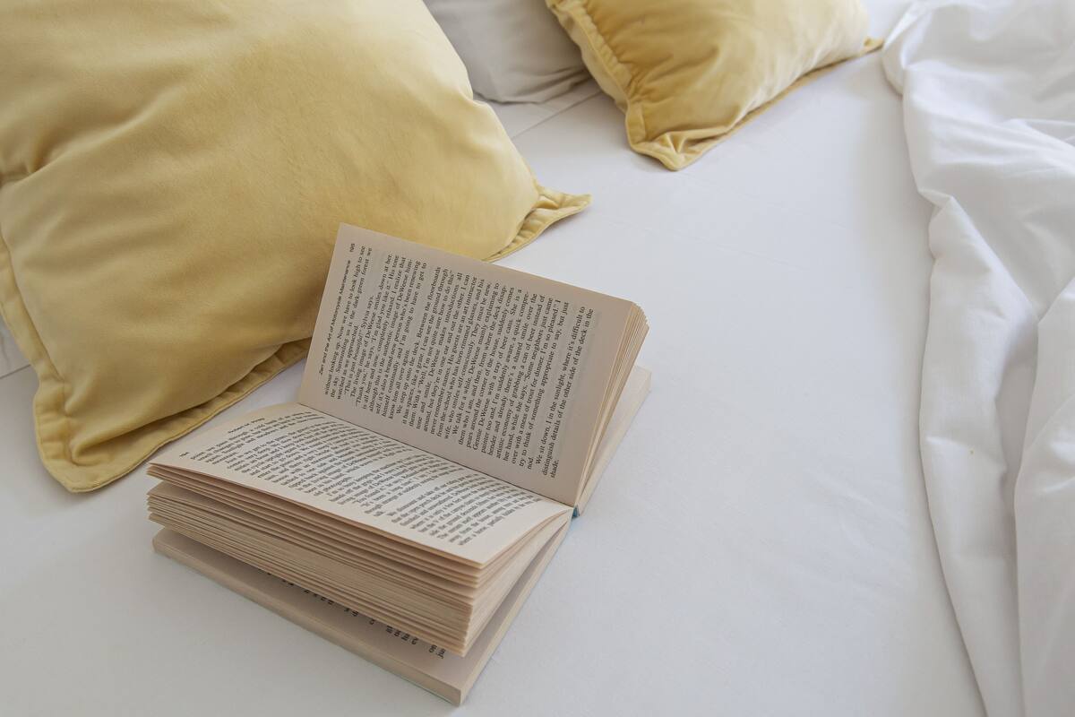 A bed with white sheets, a stack of books, and two pillows, one yellow and one white.
