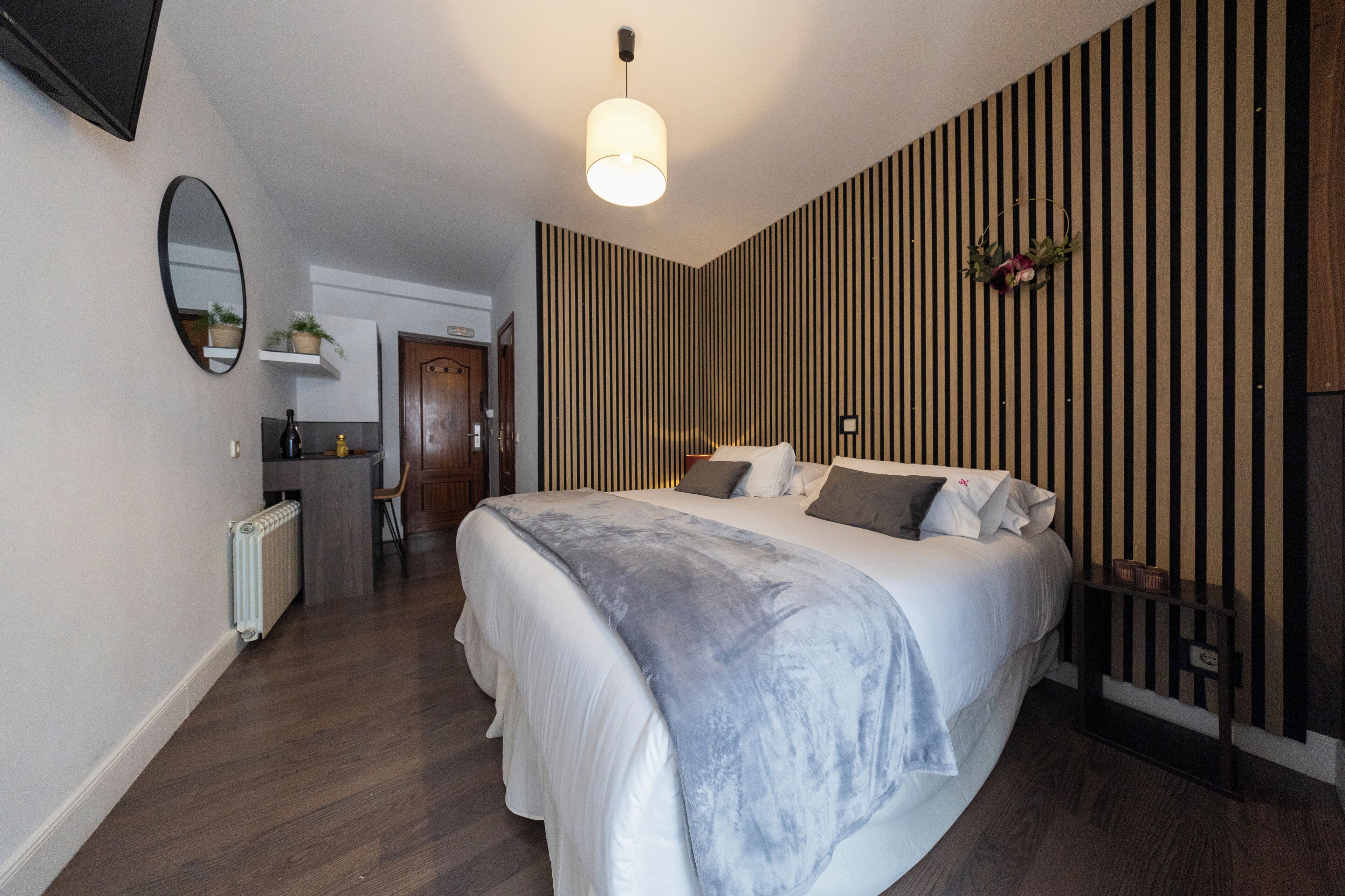 A bedroom with a bed, a bedside table, a lamp, a mirror, a radiator, and a wooden floor.