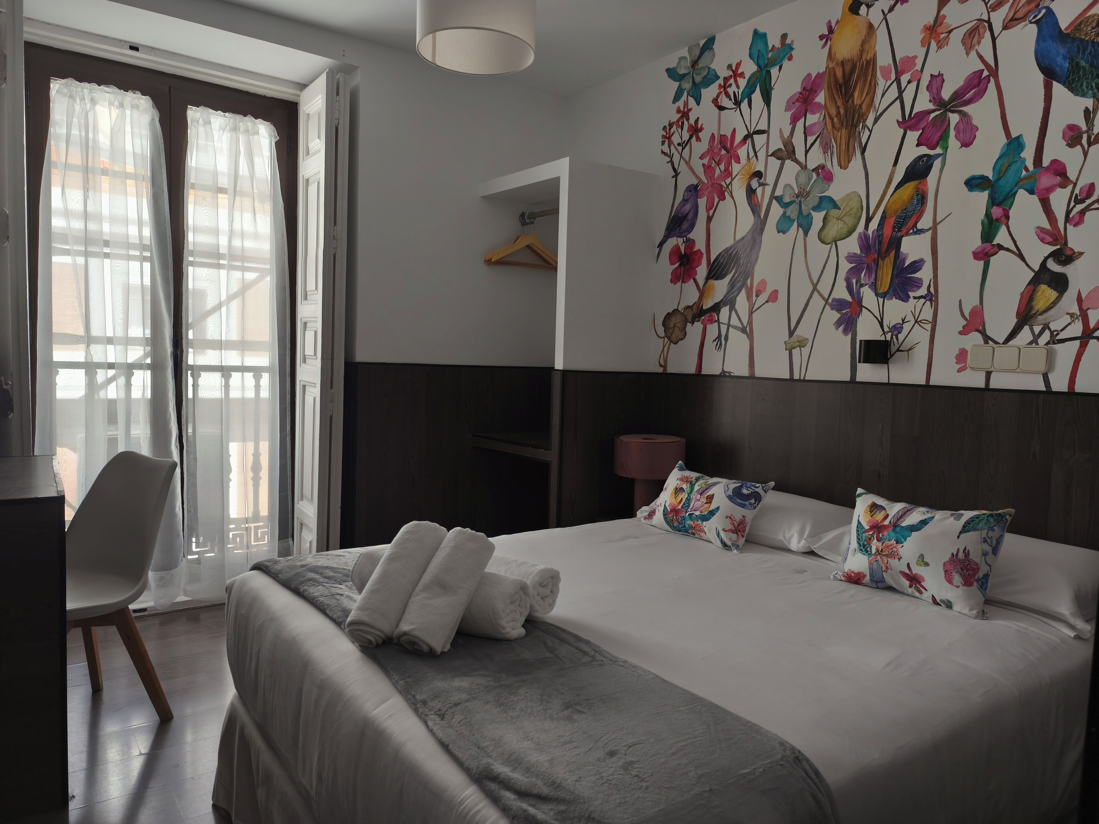 Ett hotellrum med en säng, en stol, ett skrivbord och en vägg prydd med en blommig väggmålning.