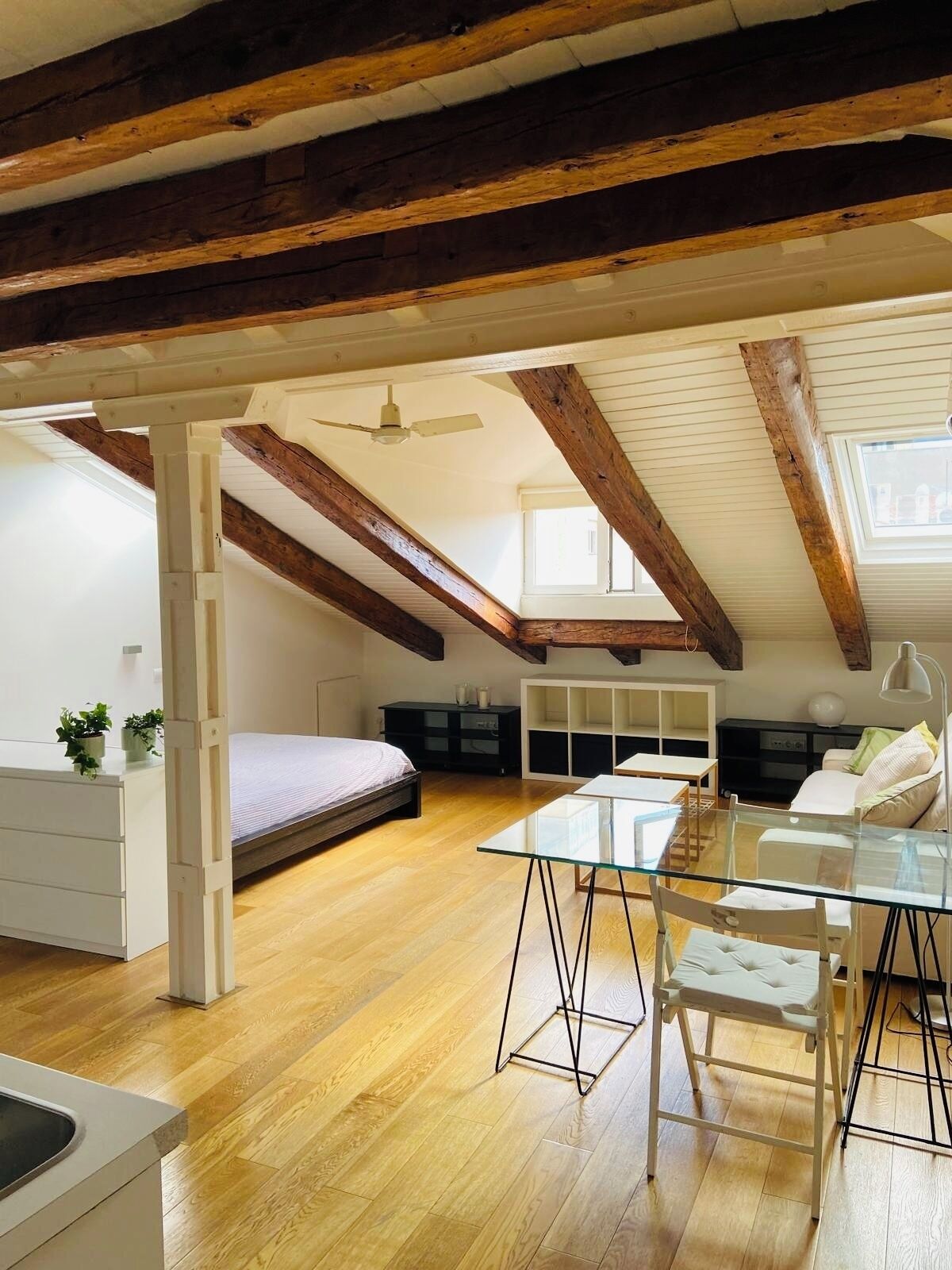 Un dormitorio en el ático con vigas de madera a la vista, una cama, una mesa de comedor de vidrio y un ventilador de techo.