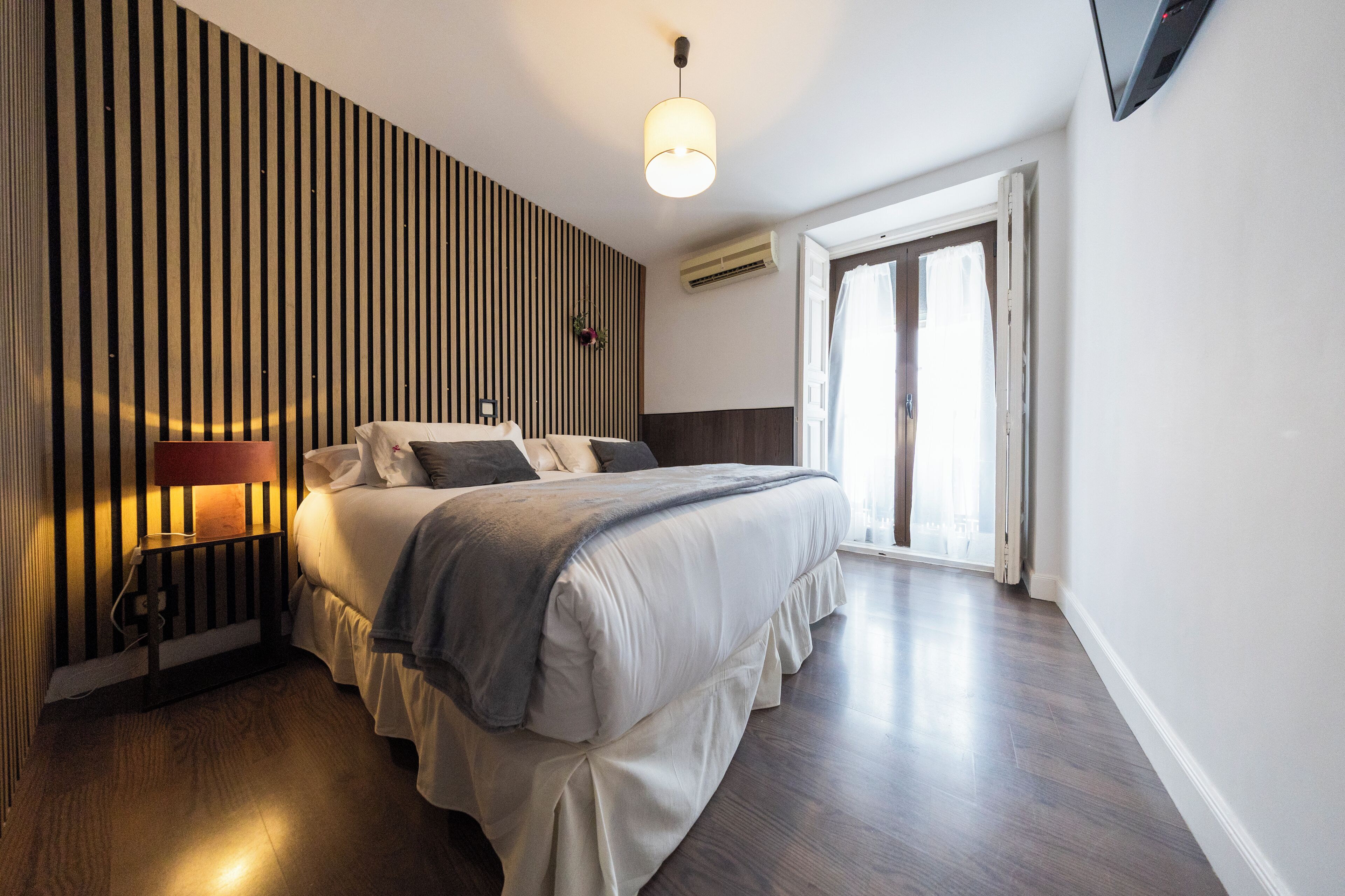 A modern hotel room with a large bed, a bedside table with a lamp, a wall-mounted air conditioner, and a sliding glass door leading to a balcony.