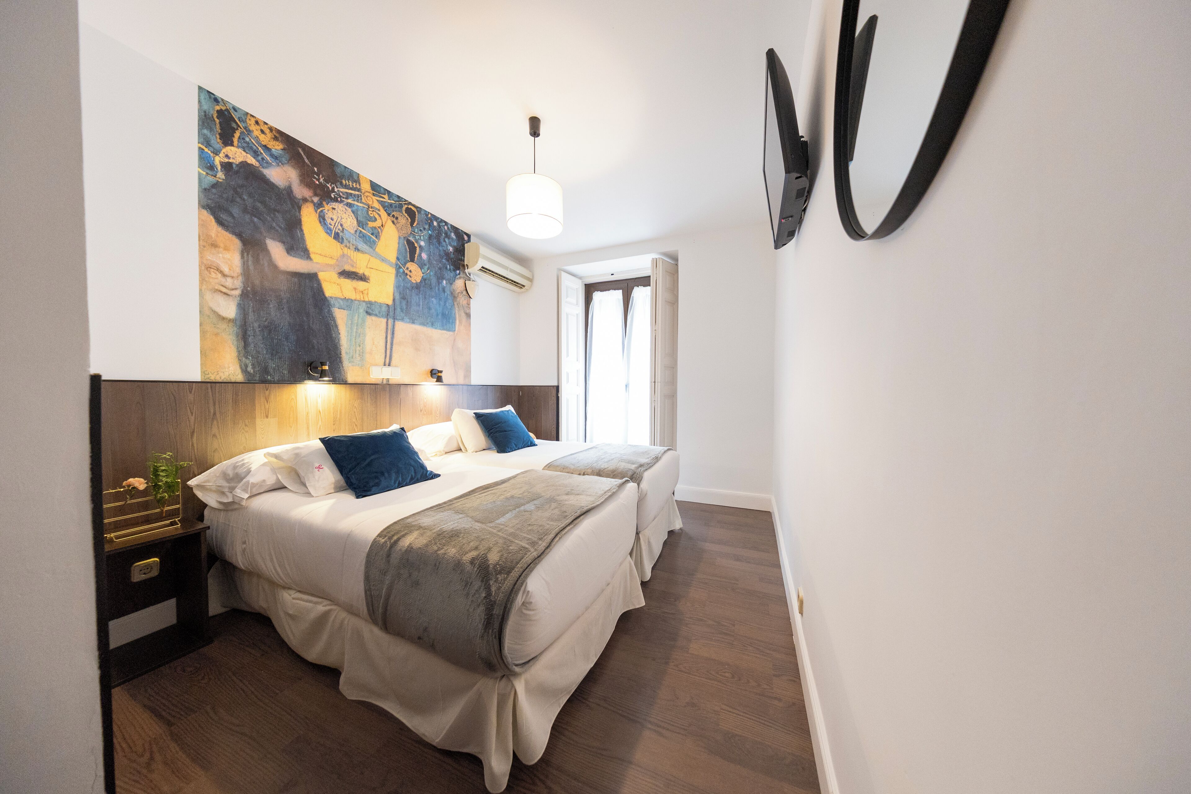 A bedroom with two beds, a painting on the wall, a ceiling light, and a door leading to another room.