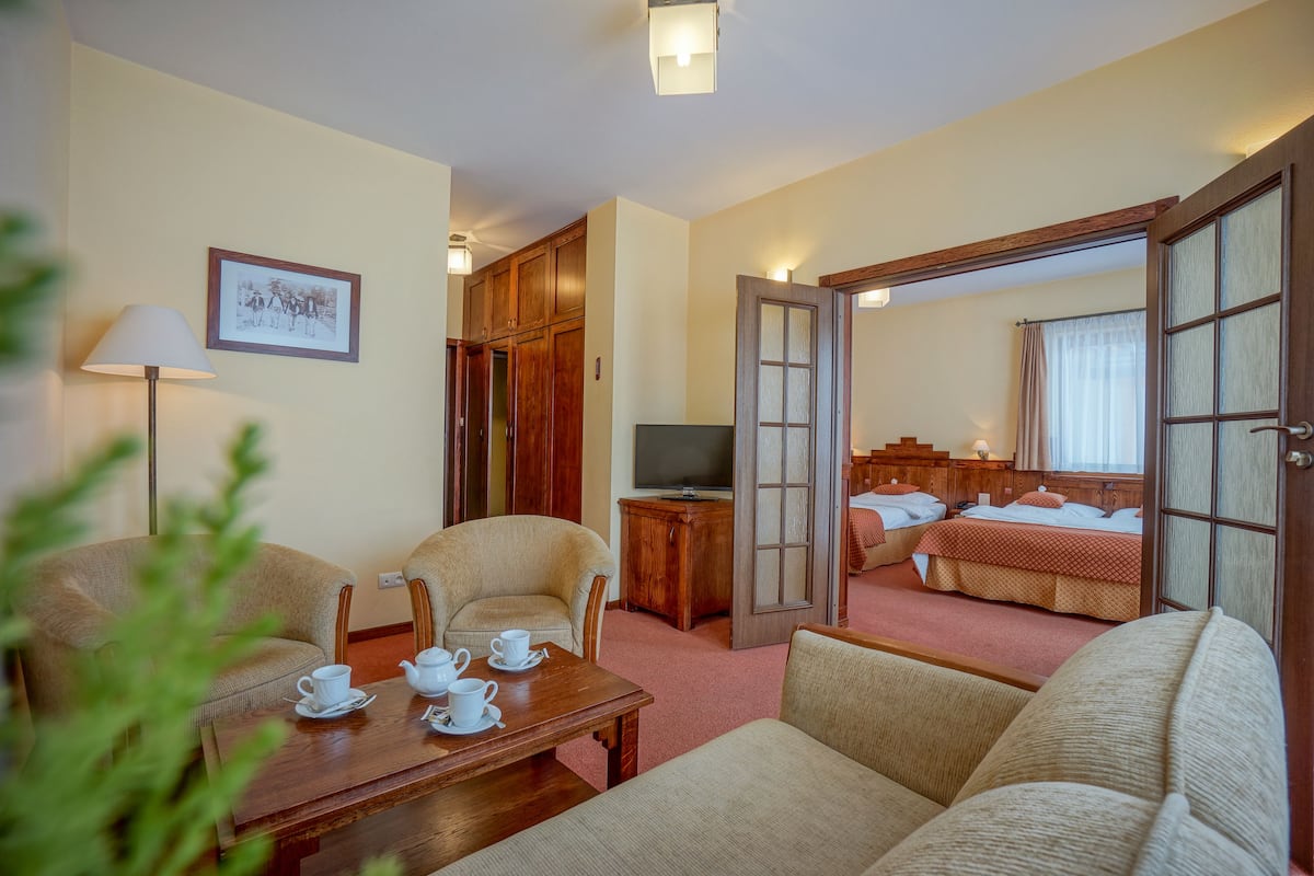 A hotel room with a wooden table set for tea, a sofa, a bed with a red bedspread, and a television on a wooden cabinet.