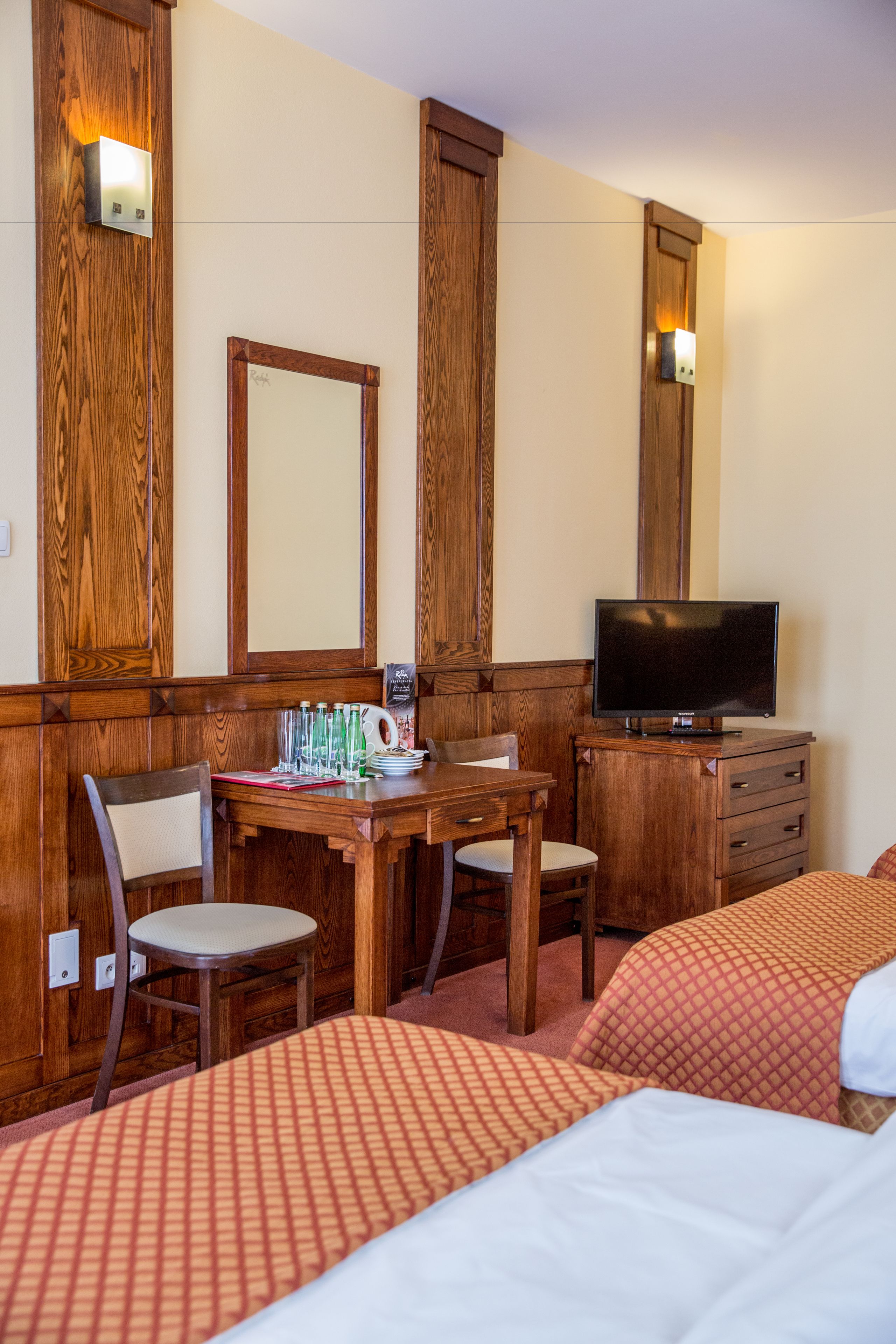A hotel room with wooden paneling, a desk with chairs, a television, and two beds with patterned bedding.