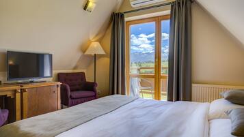 Hotelkamer met een bed, een televisie, een stoel, een lamp en uitzicht op bergen door een groot raam.
