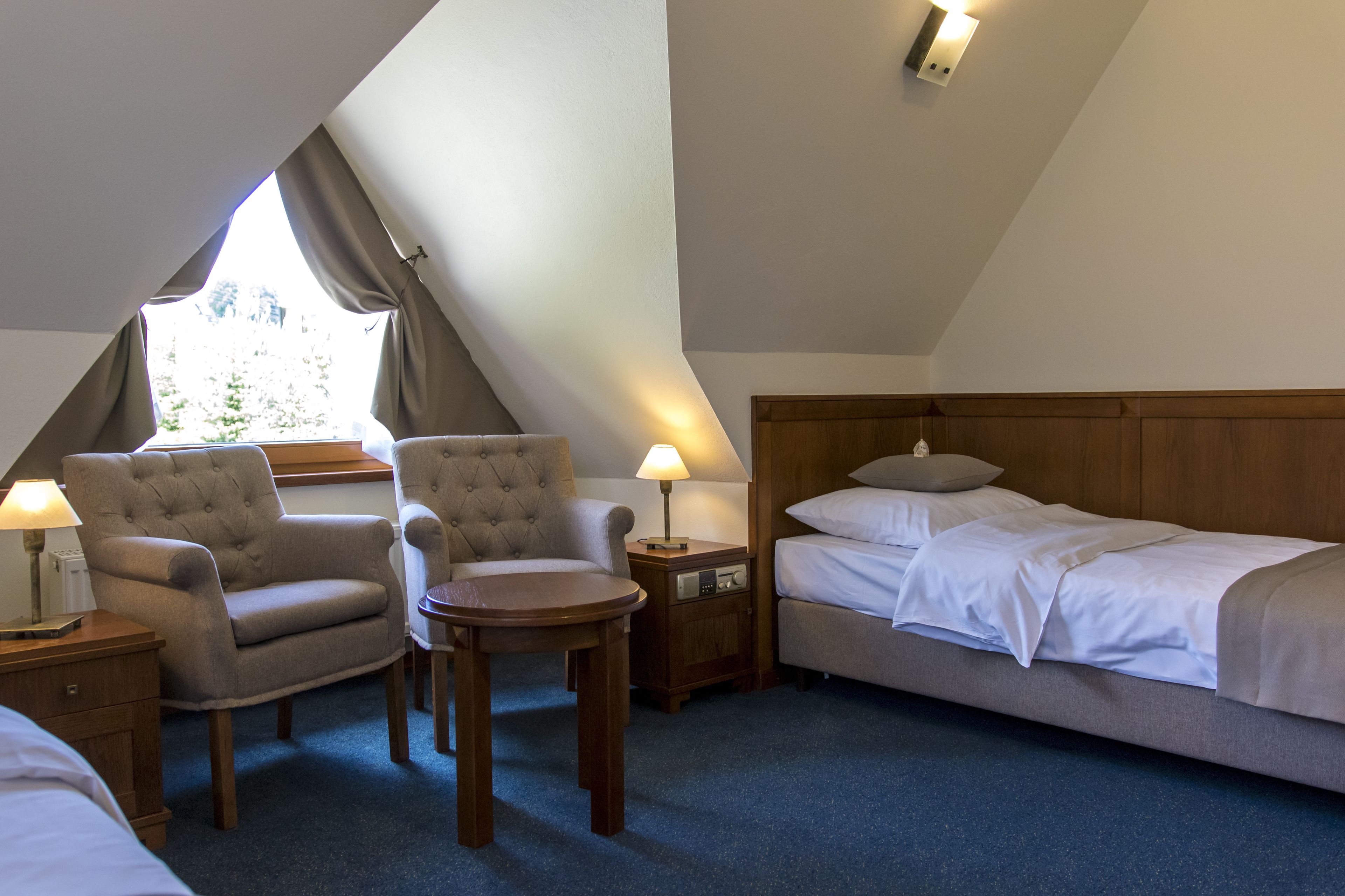 Ein Schlafzimmer mit einem Bett, zwei Sesseln, einem kleinen Tisch, einem Nachttisch mit Lampe und einem Fenster mit Vorhängen.