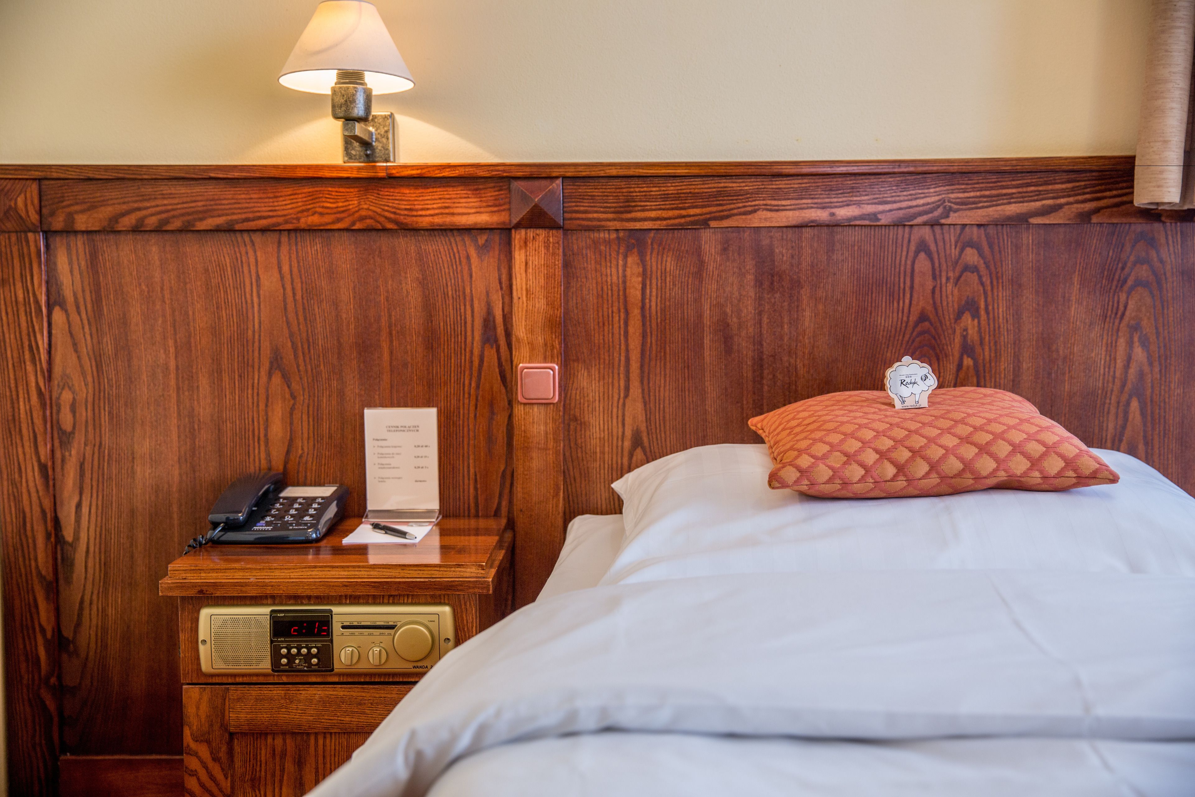 A wooden headboard with a lamp, a bedside table with a phone, a radio, and a pillow.