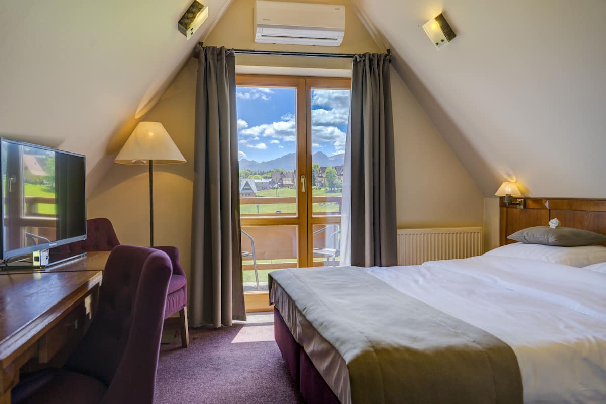 A hotel room with a bed, a desk with a television, a chair, a lamp, and a view of the outdoors through a large window.