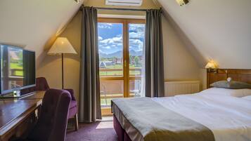 A hotel room with a bed, a desk with a television, a chair, a lamp, and a view of the outdoors through a large window.