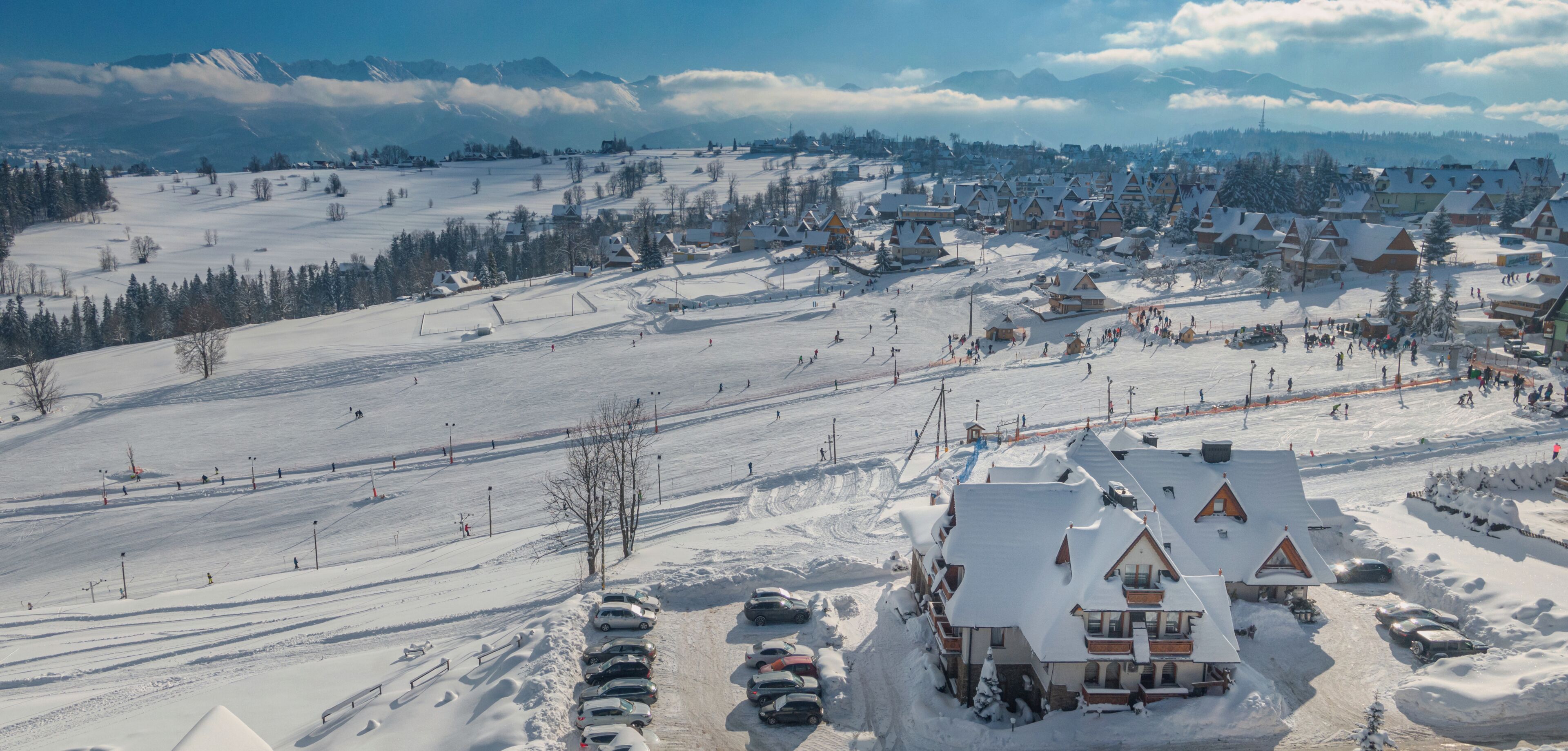 Estación de esquí nevada con edificios de estilo chalete, una pista de esquí y un área de estacionamiento.