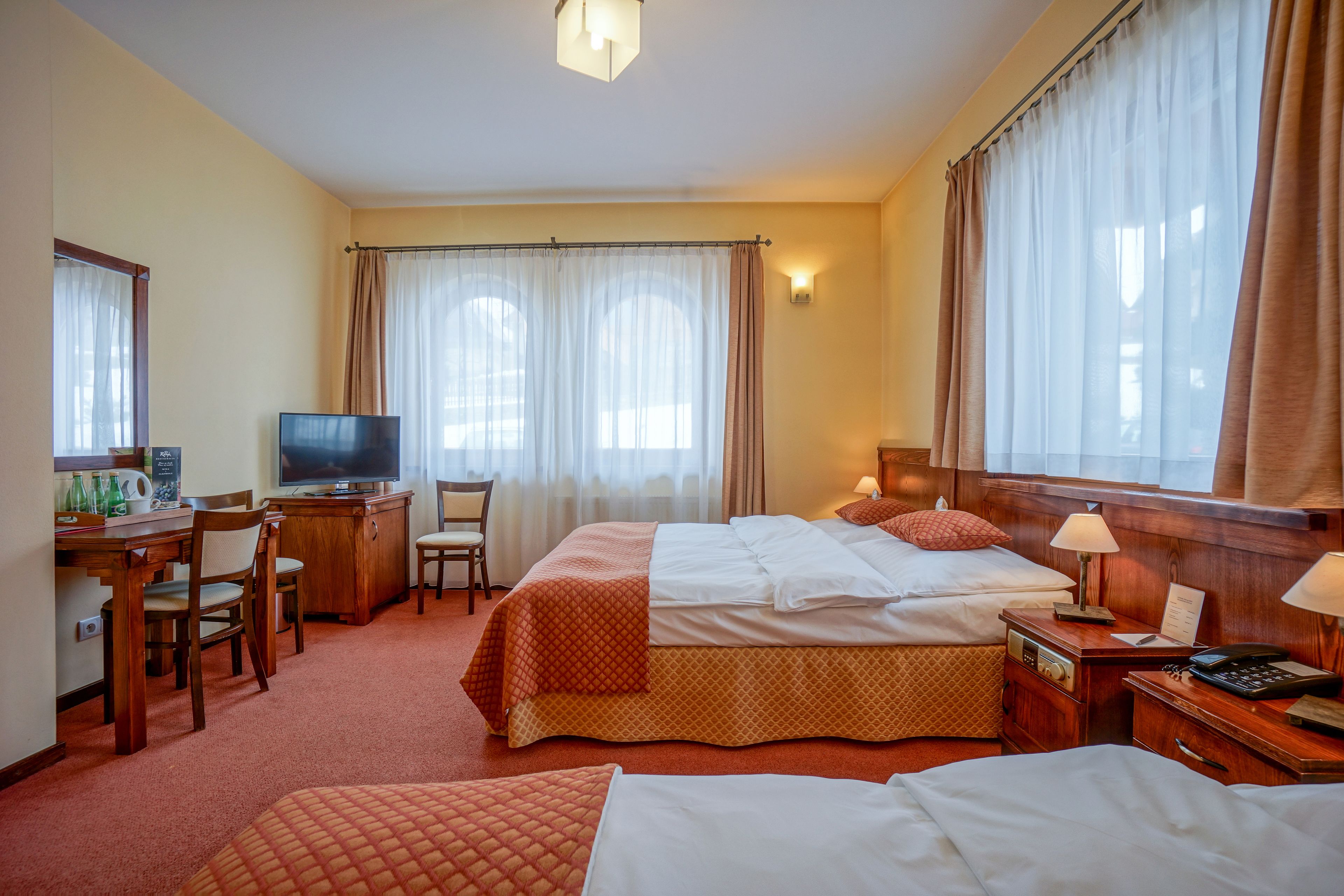 A hotel room with two beds, a desk with a television, a chair, and a small table with a lamp.