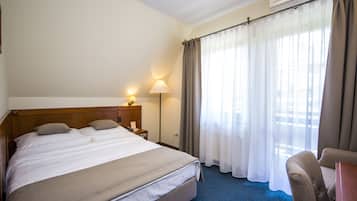 Habitación de hotel con una cama grande, una mesita de noche con lámpara, una silla y una ventana con cortinas.
