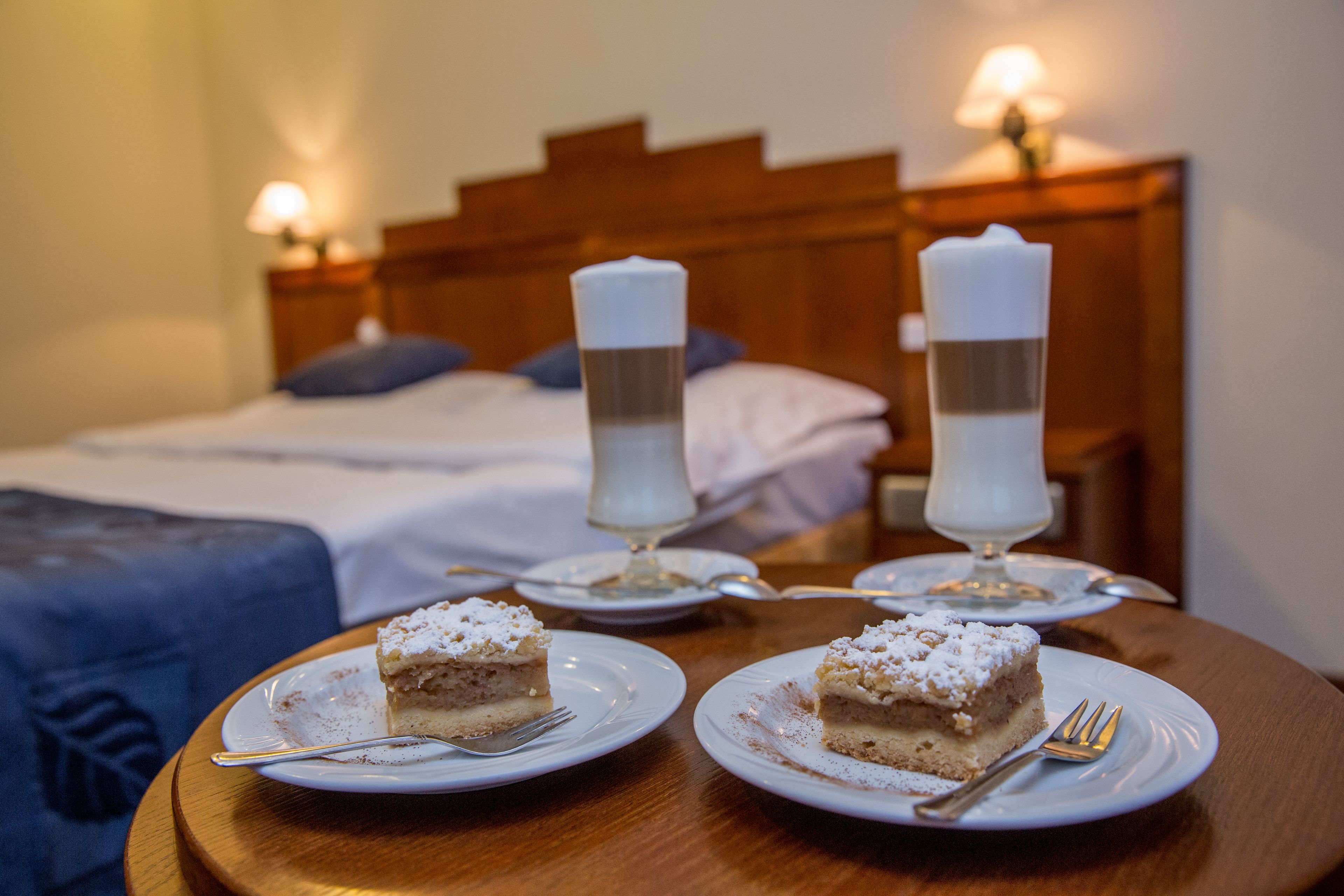 A hotel room with a neatly made bed, two white candles on bedside tables, and two plates of cake on a wooden table.