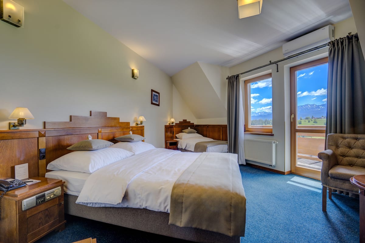 A hotel room with two beds, a wooden headboard, bedside lamps, a chair, and a view of mountains through the window.