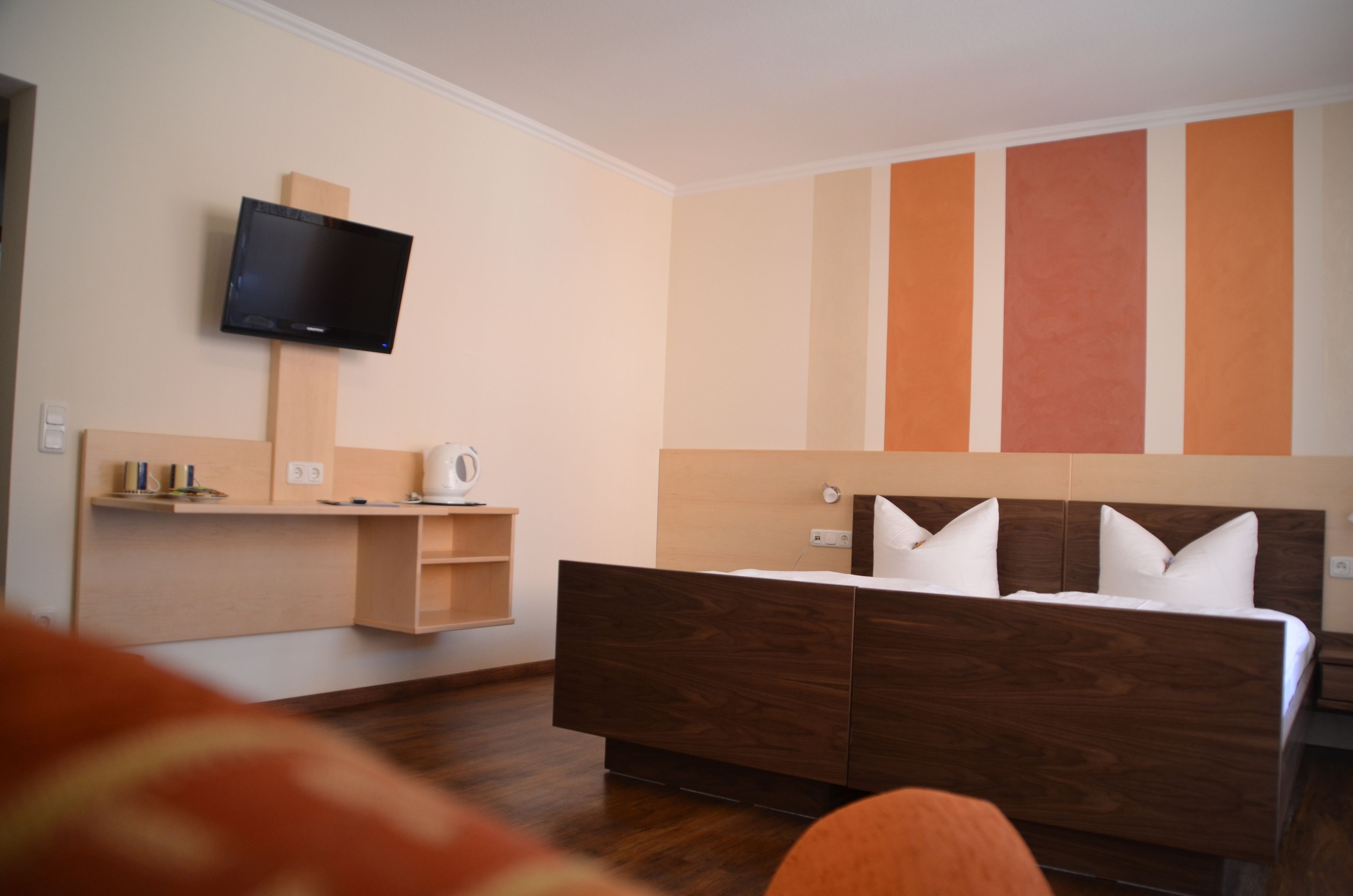 A modern hotel room with a wooden bed, a flat-screen TV mounted on the wall, and a built-in shelf with a kettle.