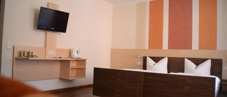 A modern hotel room with a wooden bed, a flat-screen TV mounted on the wall, and a built-in shelf with a kettle.