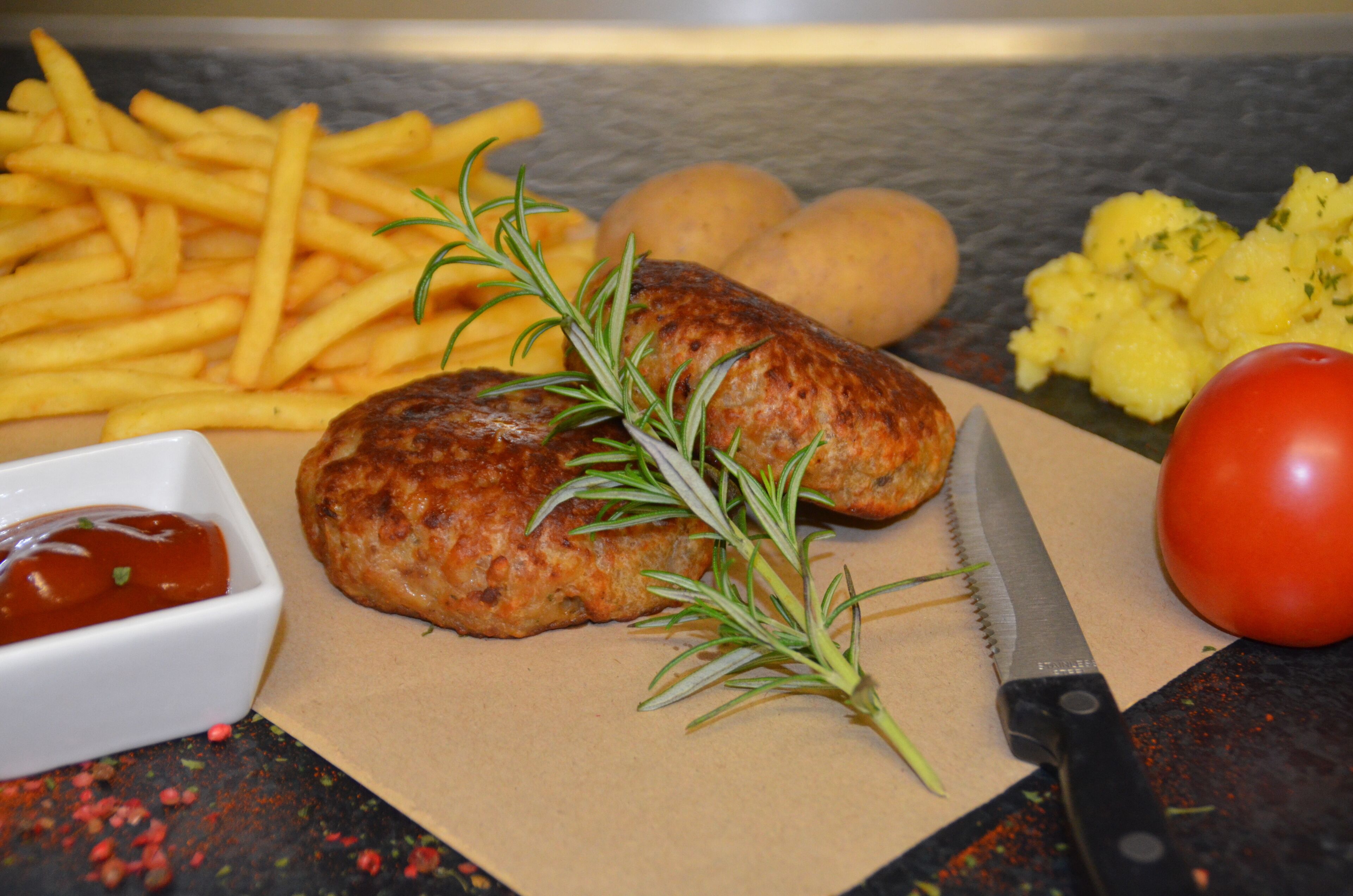 En måltid bestående av en korv, pommes frites, potatis, blomkål och en tomat, serverat på en skärbräda.