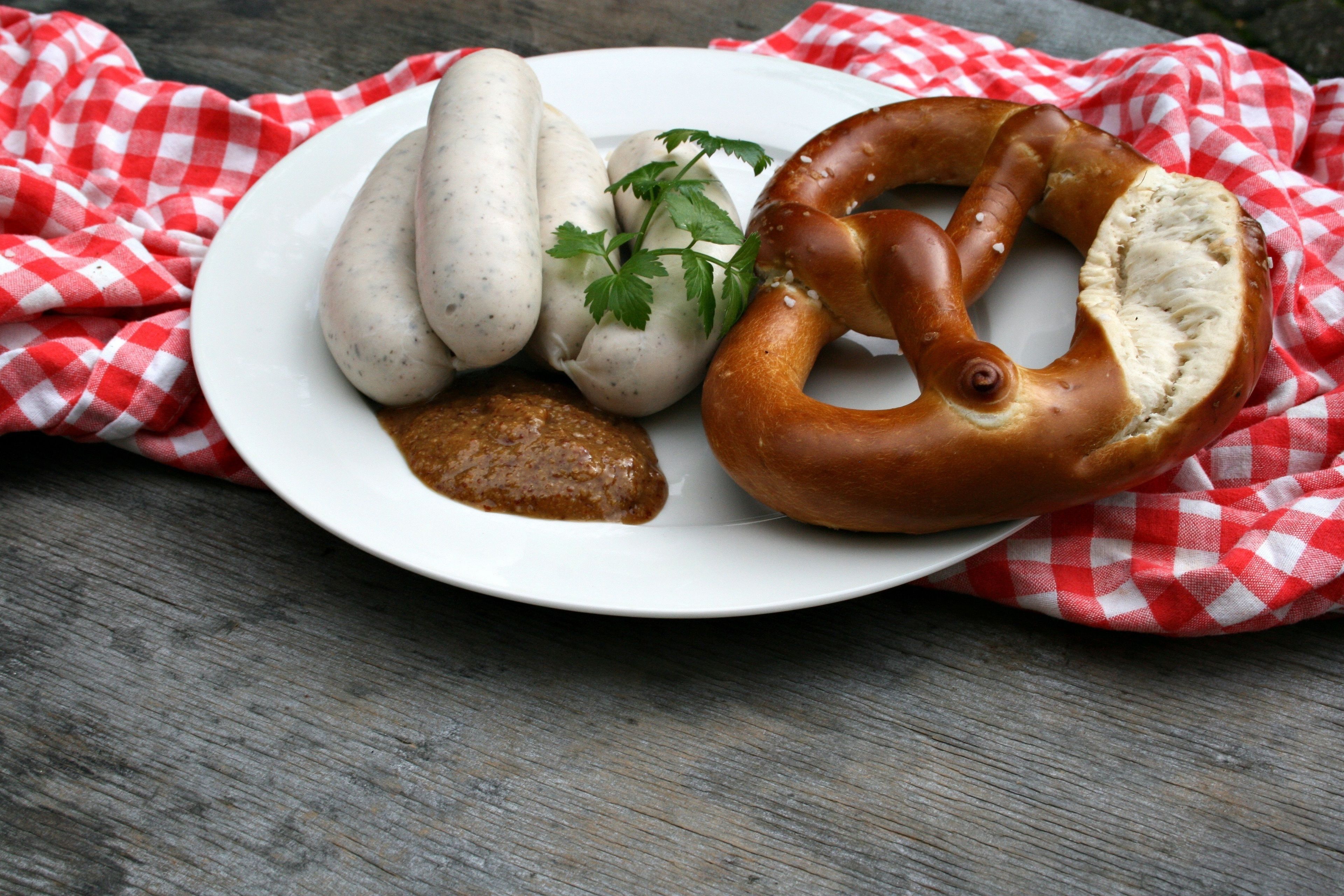 A plate with sausages, a pretzel, and a sauce on a wooden table with a red and white checkered cloth.