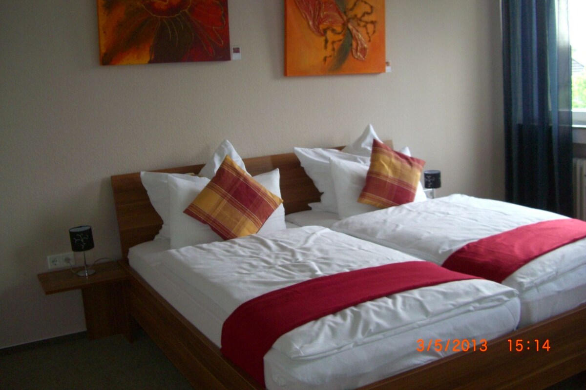 A double bed with white linens, two red and yellow pillows, and a red throw blanket. There are two paintings on the wall above the bed.