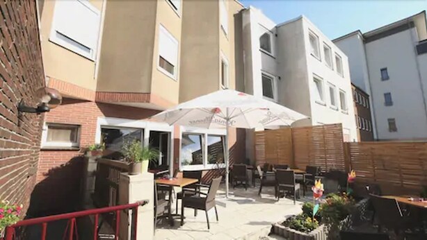 An outdoor patio area with tables and chairs under a white umbrella, surrounded by a brick wall and wooden fence. - Art Inn Hotel Dinslaken (Dinslaken)