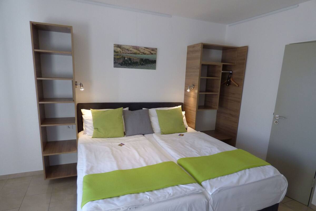 Una cama bien hecha con ropa de cama verde y blanca, un cabecero con estanterías integradas y un pequeño cuadro de paisaje en la pared.